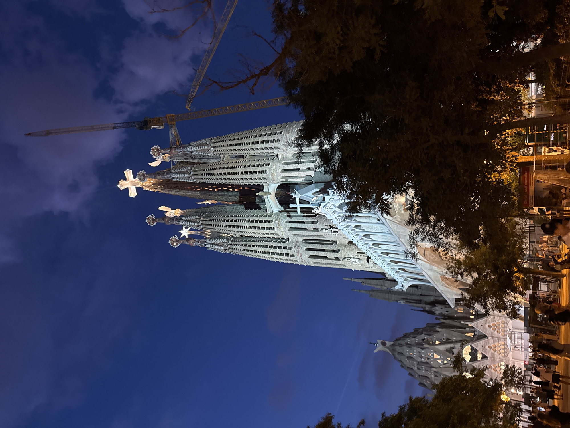 This is an exterior, eye-level, nighttime, vertical shot of the Sagrada Familia in Barcelona, Spain. The iconic basilica is illuminated, showcasing its intricate spires and facades, with a large crane positioned behind the structure, indicating ongoing construction. The sky above is a deep blue, with some clouds visible. In the foreground, silhouetted trees frame the view, and a crowd of people are visible in the lower portion of the image, many appearing to be tourists taking in the sight. The overall scene conveys a sense of awe and grandeur, with the basilica's illuminated architecture contrasting with the darkening sky and the presence of modern construction equipment.