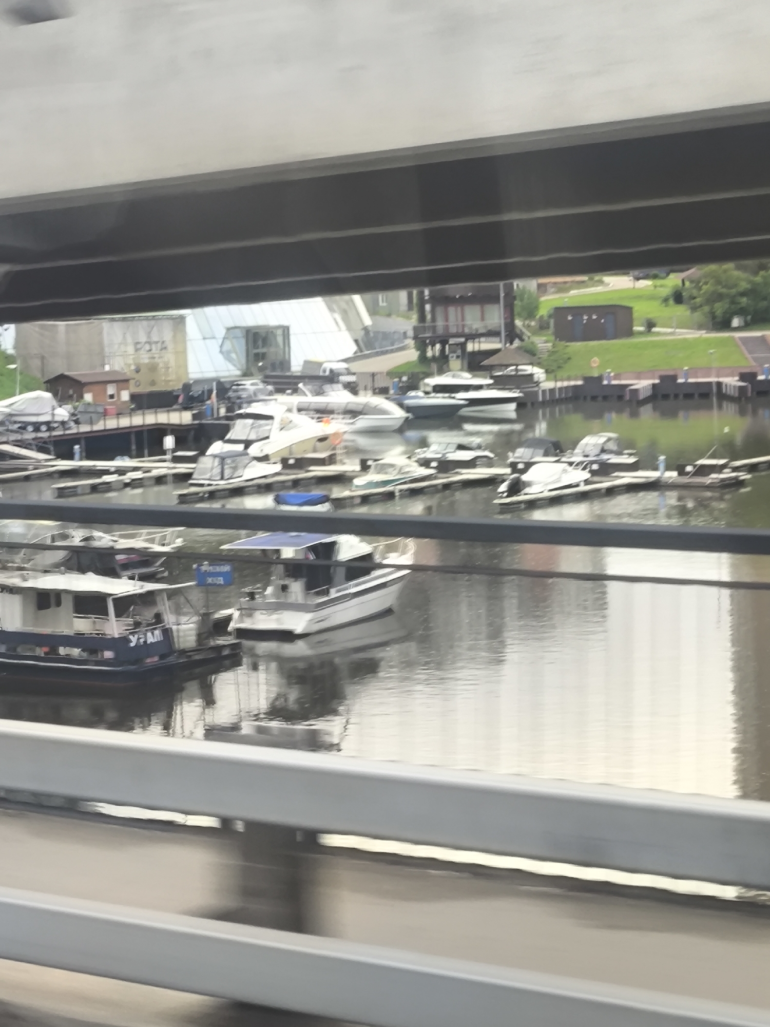 The image shows a marina filled with various boats and yachts, viewed through the structural framing of a bridge or overpass. The location is the city of Unknown, Unknown. The water is calm, reflecting the boats and surrounding structures. In the background, there are buildings and greenery. The perspective suggests the photo was taken from a moving vehicle on the bridge.
