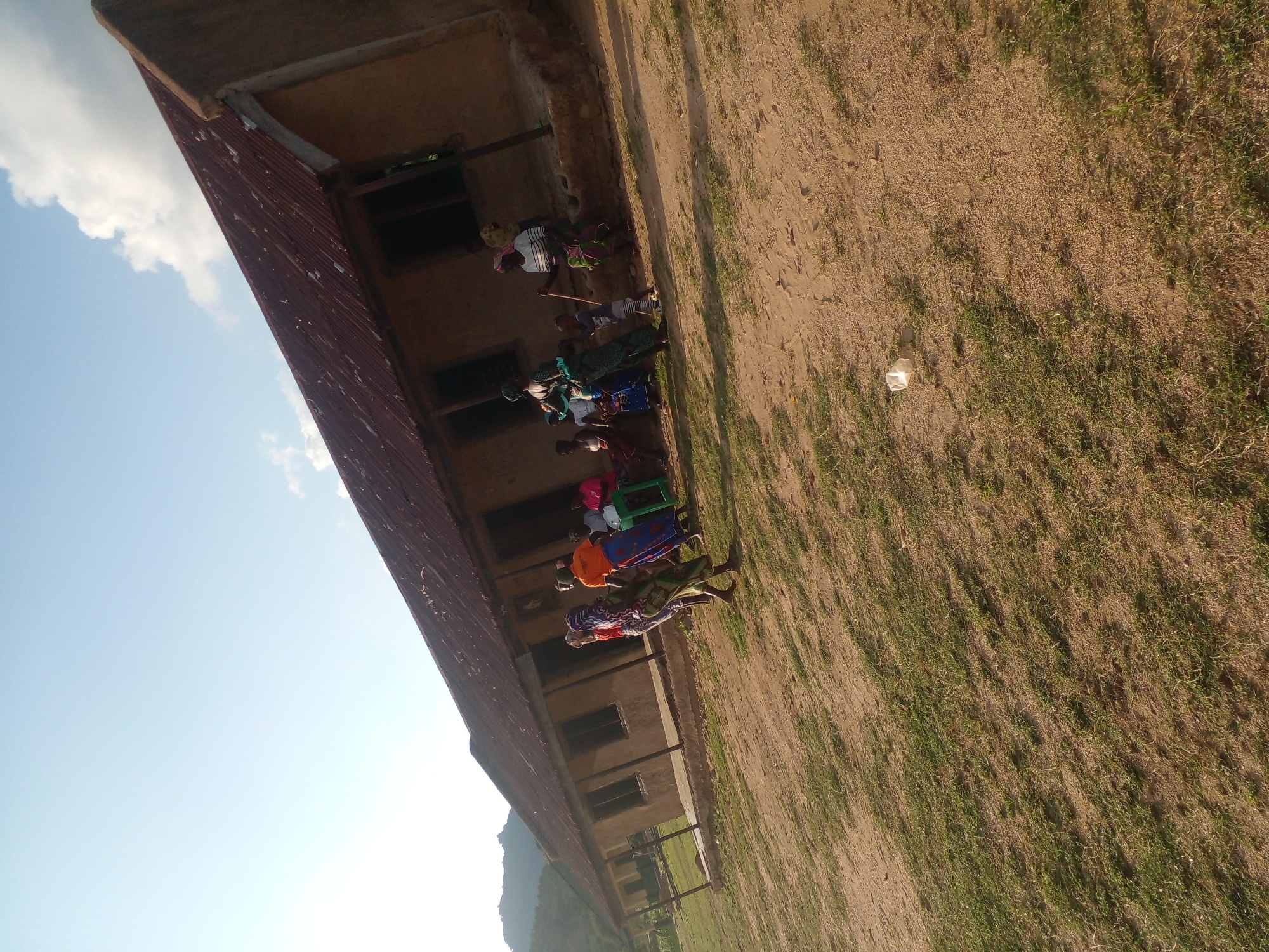 The image depicts an outdoor scene in front of a long, single-story building with a corrugated metal roof. The building appears to be constructed of mud or adobe with visible wooden support beams under the roof overhang. Several windows are visible along the building's facade. A group of people, primarily women and children, are gathered outside. Some women are wearing brightly colored wraps. One woman is standing and appears to be talking to another woman who is seated. A child is standing near a woman holding a stick. The ground in front of the building is a mixture of dirt and sparse grass, with some areas of lighter colored sand. In the foreground, a crumpled white plastic bag lies on the ground. In the background, a hilly, green landscape is visible under a clear blue sky with scattered clouds. The lighting suggests it is daytime, possibly late afternoon. The scene gives the impression of a rural or semi-rural community setting.