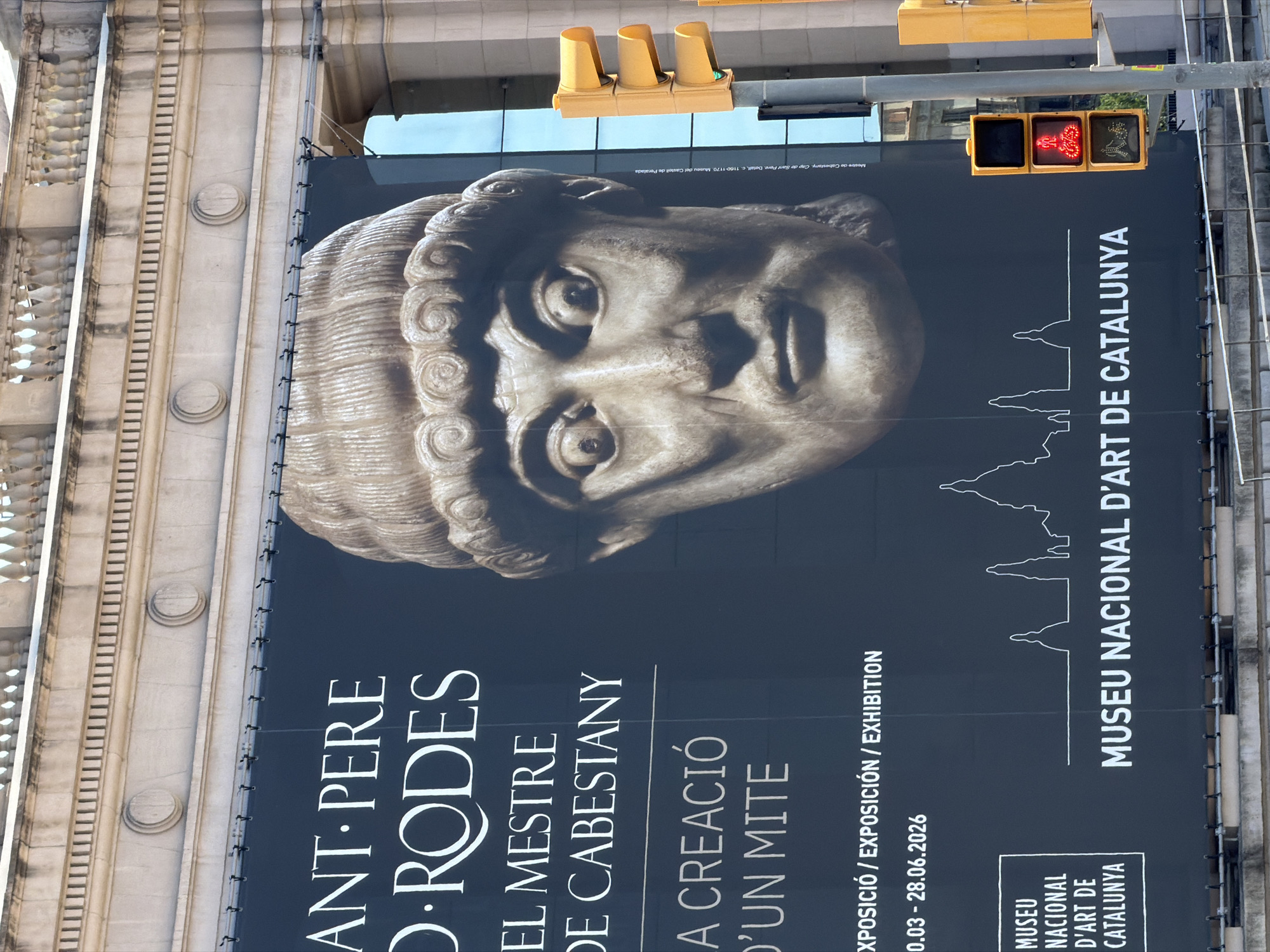 This image captures a large outdoor advertisement for an exhibition at the Museu Nacional d'Art de Catalunya in Barcelona, Spain. The advertisement features a close-up of a stone bust of a man with distinctive curly hair and wide eyes, against a dark background. Prominently displayed are text elements in white, including the exhibition title "ANT·PERE D·RODES EL MESTRE DE CABESTANY" and "A CREACIÓ D'UN MITE". Below this, exhibition dates and further text including "EXPOSICIÓ / EXPOSICIÓN / EXHIBITION" and "0.03 - 28.06.2026" are visible. The logo for "MUSEU NACIONAL D'ART DE CATALUNYA" is in the bottom left corner, alongside a stylized outline of Barcelona's skyline. To the right of the advertisement, a traffic light pole with three yellow signal heads and pedestrian signals is visible. The top of a building with classical architectural elements, including columns and balustrades, forms the backdrop. The scene appears to be outdoors, likely on a sunny day, given the lighting.