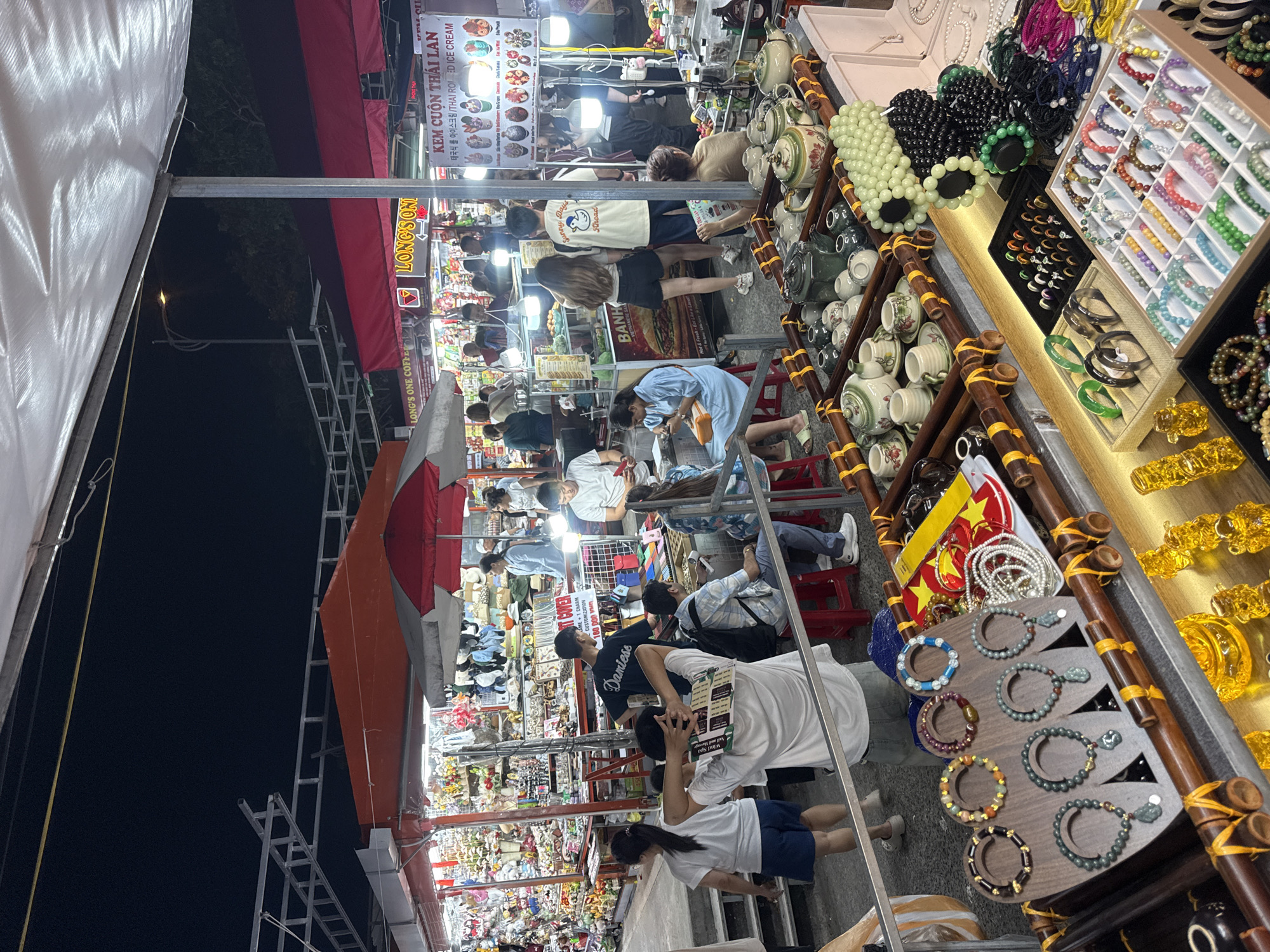 Here is a description of the image:
The image depicts a bustling street market at night in an unknown city, filled with people browsing various stalls. The market is illuminated by bright lights, creating a vibrant atmosphere.
Stalls selling food items like "KEM CUON THAI LAN" (Thai Rolled Ice Cream) and "BANH" (likely referring to Vietnamese sandwiches or snacks) are visible. A wide array of merchandise is on display, including jewelry, teapots, souvenirs, and clothing.
People are walking through the market, examining goods, and interacting with vendors. Some are wearing masks. The stalls are covered with awnings, some of which have advertisements or logos. The image captures a lively scene of a typical night market with diverse offerings and a vibrant crowd.