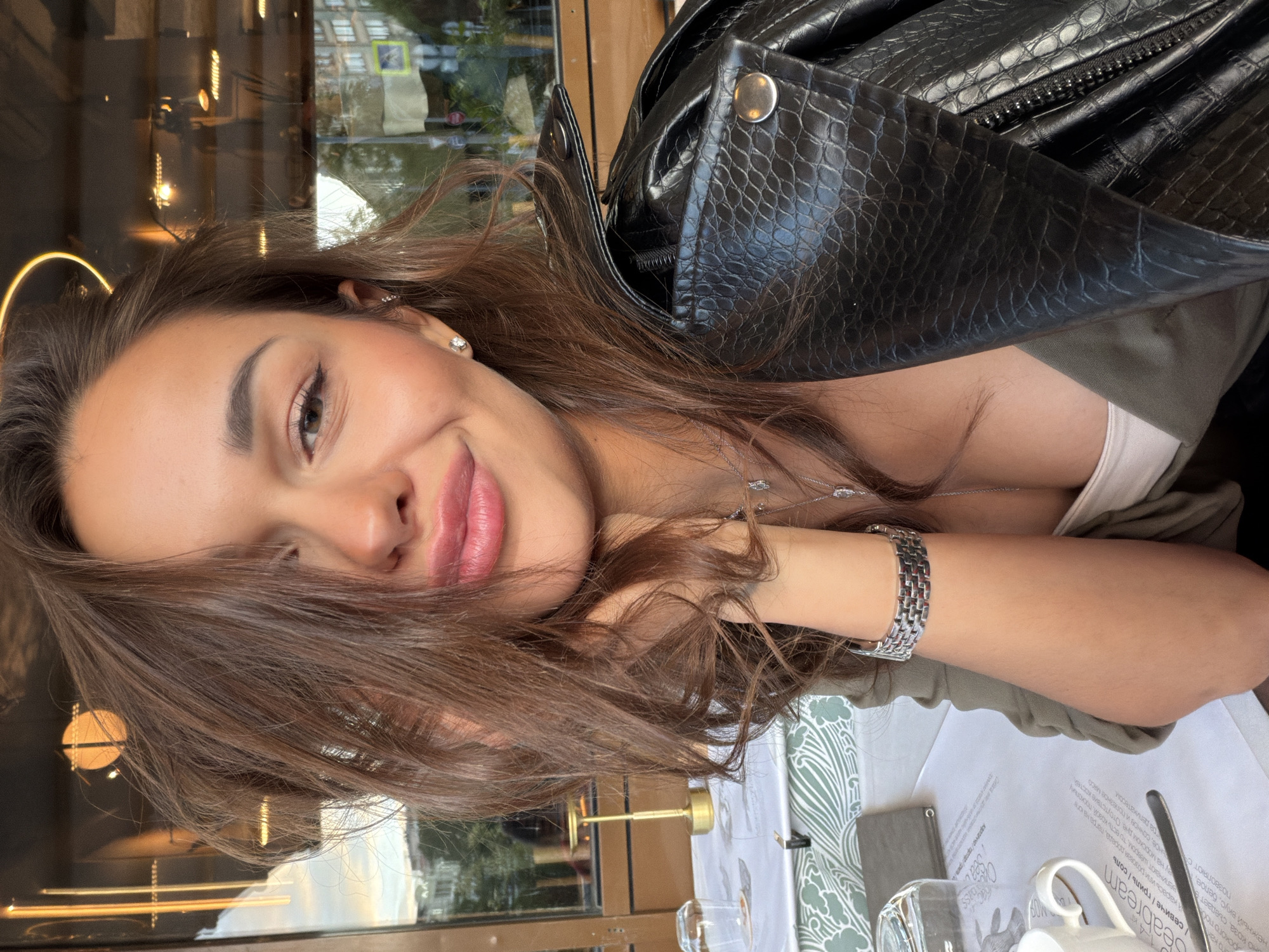 In Moscow, Russia, a woman with brown hair is captured in a selfie at a restaurant. She is wearing a dark, textured leather jacket over a white top, and a silver bracelet adorns her left wrist. Her right hand is resting on her chin, and her lips are slightly parted as she smiles at the camera. The background reveals the restaurant's interior with warm lighting and a window offering a glimpse of the city's buildings and sky. The table is set with a white tablecloth, a glass, and a menu.