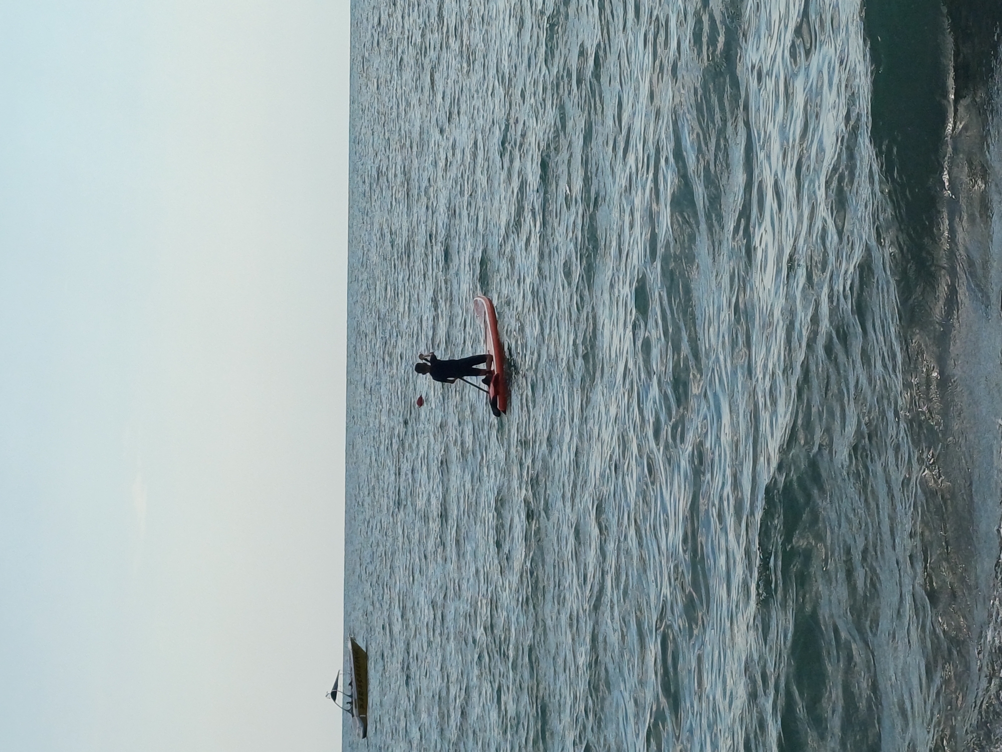 A person in a black wetsuit is paddleboarding on a calm sea. The paddleboard is red and has a white stripe down the center. The person is standing and holding a paddle, with their body angled slightly forward. In the background, there is a light blue sky and a slightly choppy sea. There is another small boat visible in the distance, to the left of the paddleboarder. The image appears to have been taken during the day, with soft lighting. The overall mood is serene and peaceful.