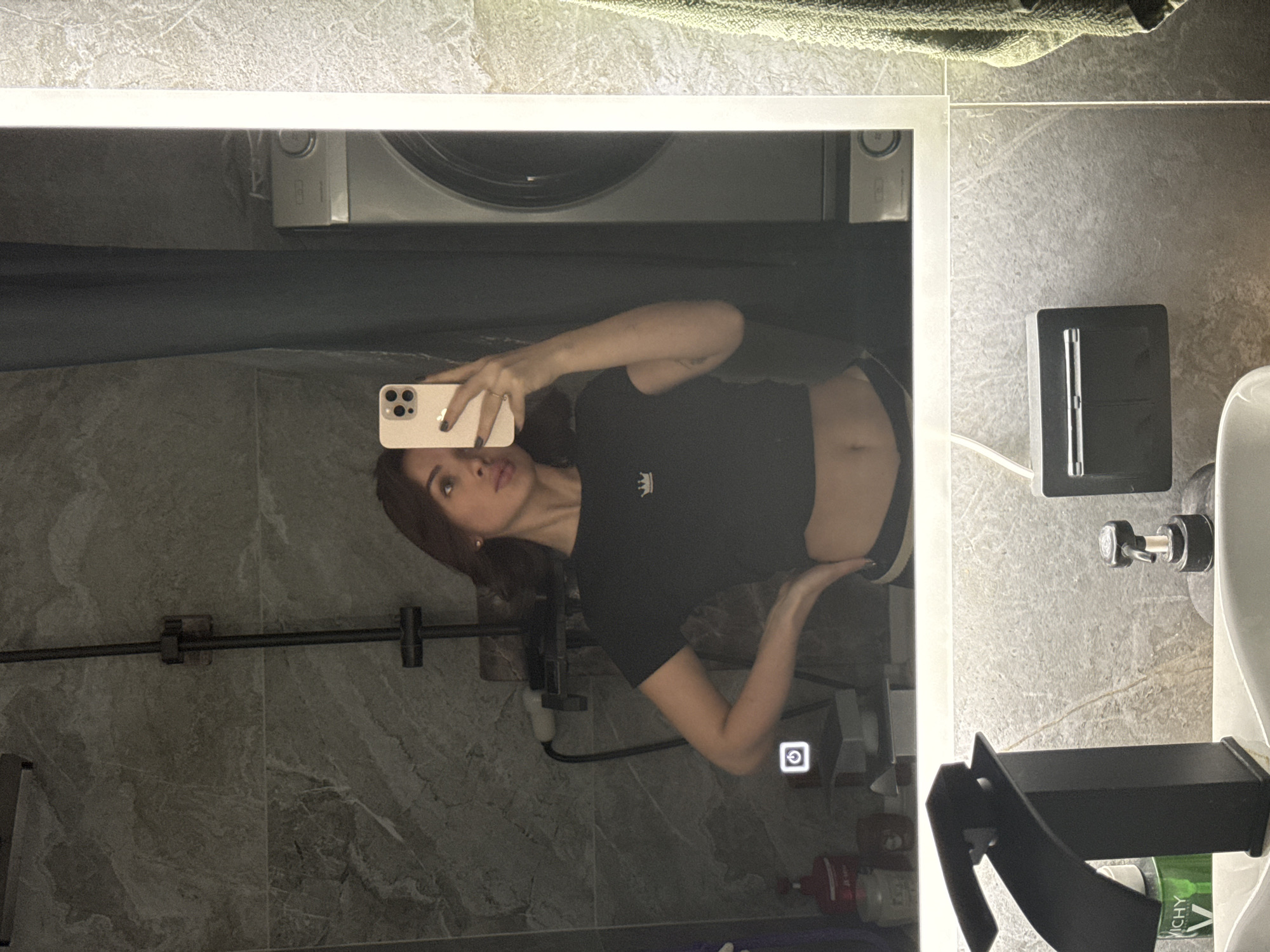 A woman is taking a selfie in a bathroom mirror. The bathroom has gray-toned tile walls and contains a washing machine mounted above the sink. The woman is wearing a dark-colored crop top with a small crown emblem. The sink, soap dispenser, and other bathroom fixtures are visible. The location may be in Лобаново, Russia.