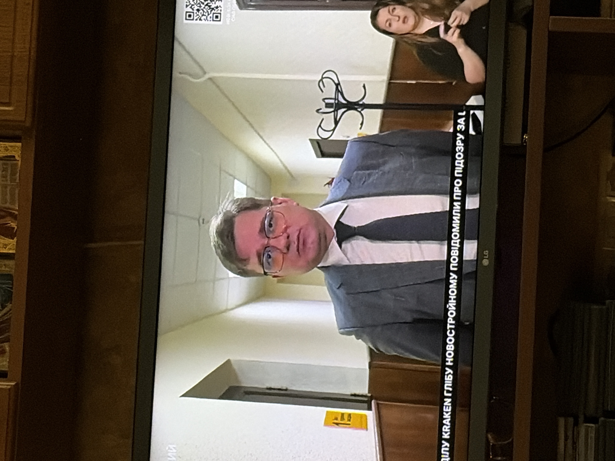 A medium shot shows a man in a suit and tie with glasses, looking forward. He is standing in a hallway with wooden paneling on the walls and a tiled floor. To his right, a woman in a black shirt is looking down and gesturing with her hands. A QR code is visible on the wall behind the man. The text on the screen reads "ДІЛУ KRAKEN ГЛІБУ НОВОСТРОЙНОМУ ПОВІДОМИЛИ ПРО ПІДОЗРУ ЗА L".