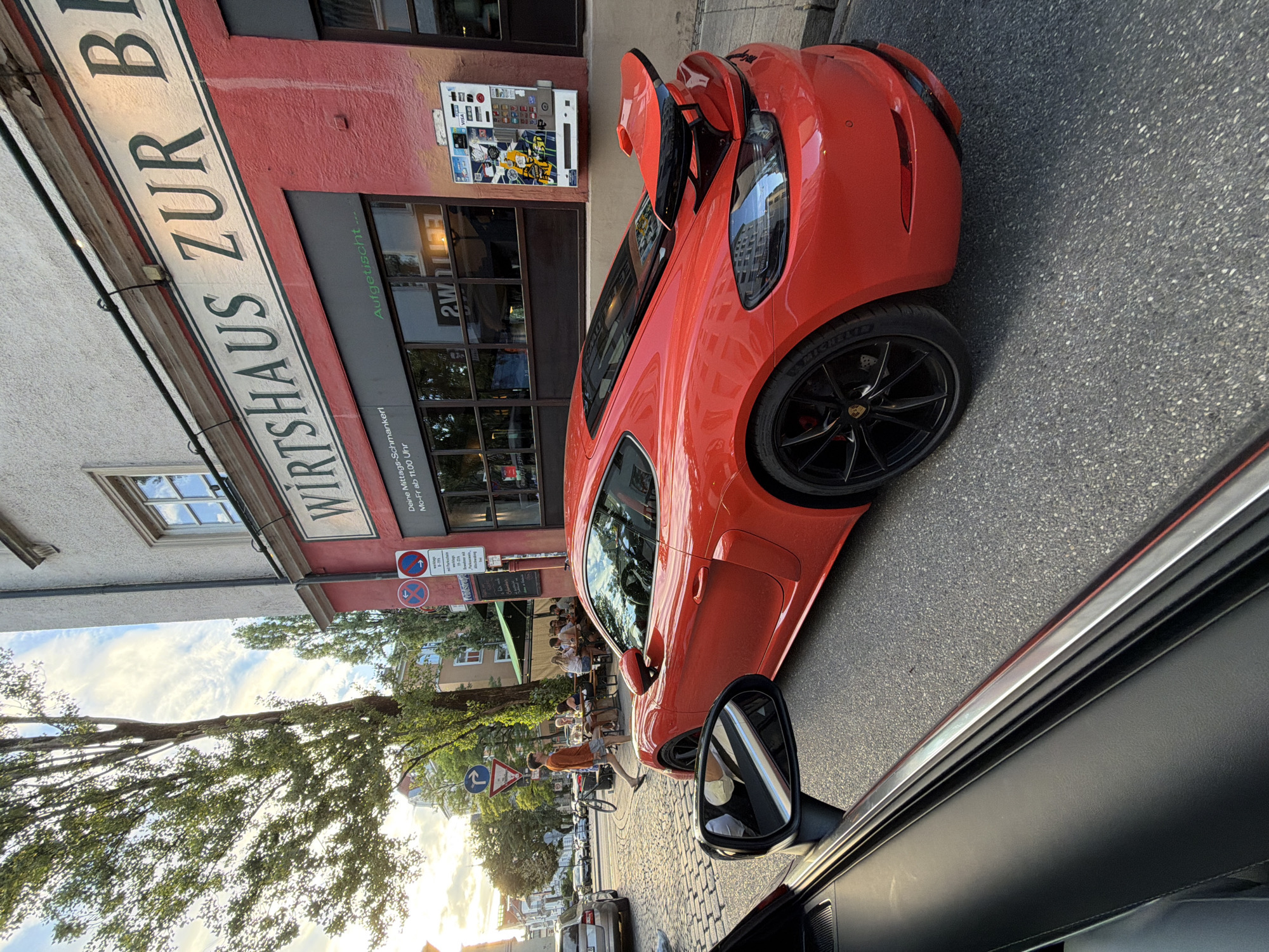 The image depicts a vibrant street scene in Munich, Germany, likely during the late afternoon or early evening given the warm lighting. In the foreground, a sleek, red Porsche sports car is parked on a cobblestone street. The car's distinctive spoiler and black wheels are visible. To the left, the interior of another vehicle is partially visible, showing a black door panel and a silver trim piece.
The background reveals a charming establishment with a red facade, bearing a large sign that reads "WIRTSHAUS ZUR BE". Beneath this sign, a dark-paneled storefront with reflective windows is visible. A small flyer or advertisement is attached to the wall to the right of the storefront. Further down the street, several people are seated at outdoor tables, enjoying a meal or drinks under a green awning. A young man in an orange shirt walks across the cobblestone street, his gaze directed elsewhere. Trees with lush green foliage line the street, and a bicycle is parked near the outdoor seating area. The sky above is a mix of blue and white clouds, indicating pleasant weather. The scene conveys a relaxed and lively urban atmosphere.