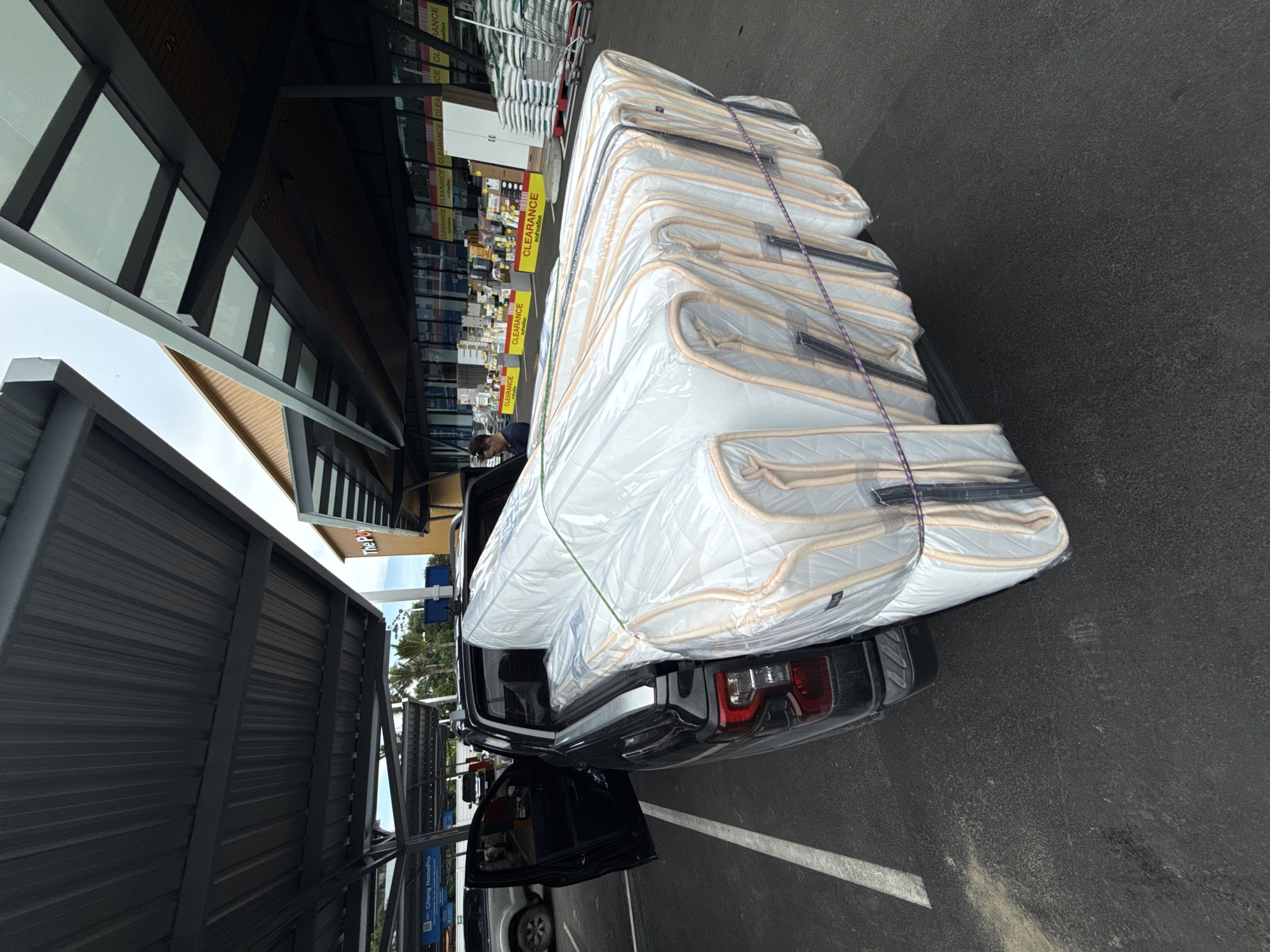 In Baan Chaweng Noi, Thailand, a black pickup truck is parked under a gray metal awning, its bed loaded with a stack of mattresses wrapped in clear plastic. The mattresses are secured with green and purple ropes. The truck's driver-side door is open. In the background, a building with large glass windows displays "CLEARANCE" signs. Stacks of bagged materials and shopping carts are visible outside the building. A person is standing near the entrance of the building, looking towards the truck. The ground is paved asphalt.