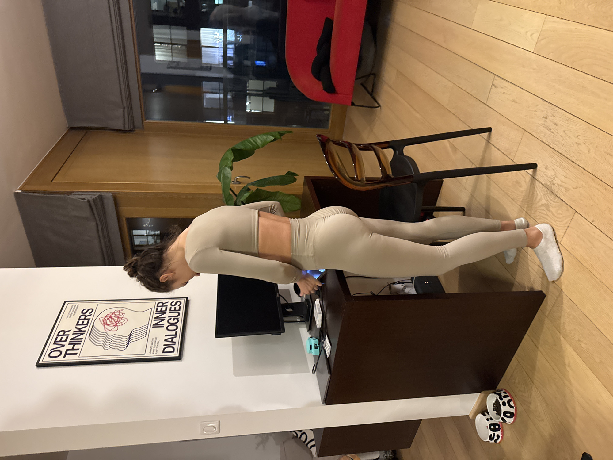 A woman in a light-colored workout outfit is performing a plank on a dark brown desk in a room with light-colored wooden floors. Her head is near the desk, and her feet are extended towards the right. A black computer monitor is on the desk. There is a framed poster on the wall to the left with the words "OVER THINKERS" and "INNER DIALOGUES." A green potted plant is behind the desk, and a wooden chair is to the right of the plant. Further back, a red chair is visible. The background shows a window with a view of a cityscape at night. Based on the architecture visible in the window, it is likely the location is within the city of Warsaw, Poland.