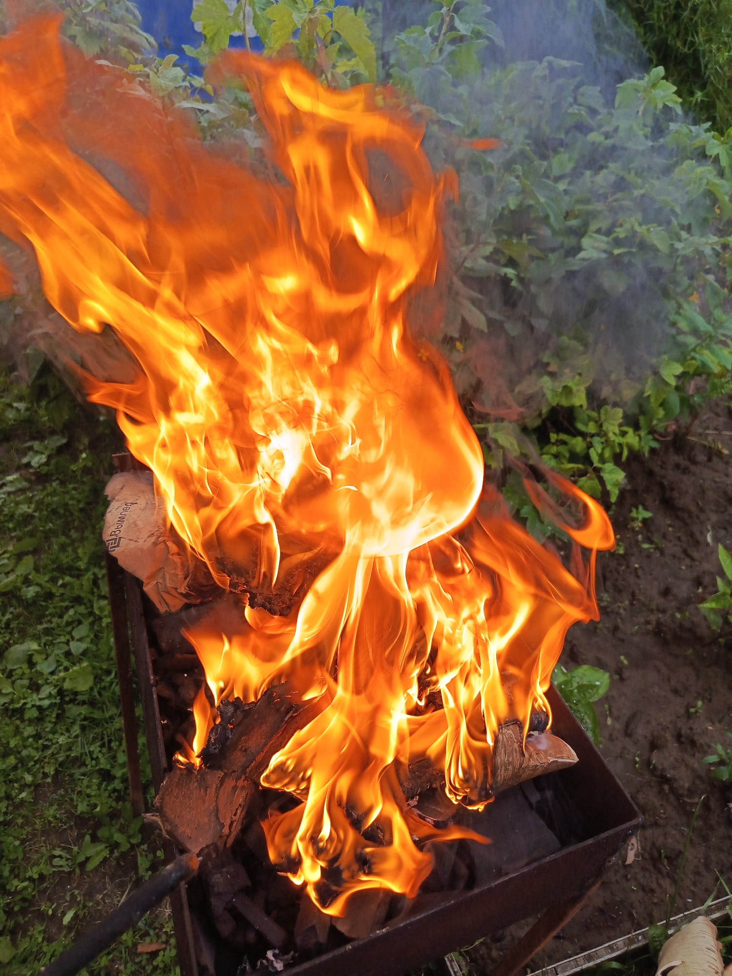 A close-up, high-angle outdoor shot captures a barbecue grill ablaze with large, bright orange flames. The fire is consuming pieces of wood and what appears to be a crumpled paper bag within the metal grill. Wisps of white smoke rise from the flames and drift to the left. The grill is situated on a patch of green grass, with brown soil and more green foliage visible in the background. The setting is natural, likely a backyard or park. No people are present in the image.