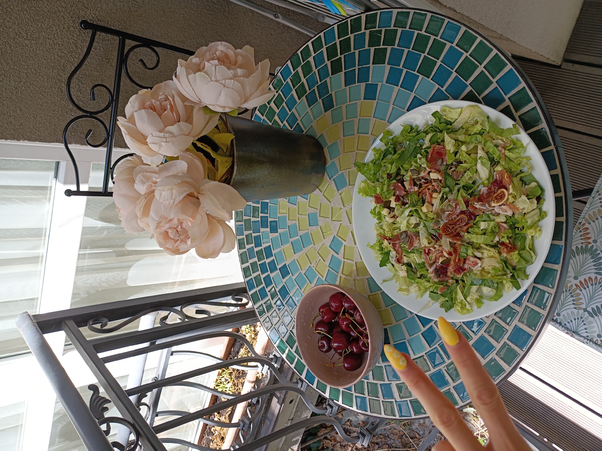 A person's hand with bright yellow nail polish is in the lower left corner of the image, holding up two fingers in a peace sign. In front of the hand, on a mosaic-tiled table, is a white plate filled with a green salad topped with thinly sliced prosciutto, cheese cubes, seeds, and possibly dried figs. To the left of the plate, a light pink bowl contains dark red cherries. Behind the table, a vase of light pink artificial flowers sits on the table. The table is set on a wooden deck, with a metal railing visible in the background. The setting appears to be an outdoor balcony or patio, possibly in Sofia, Bulgaria, given the sunlight and the architecture.