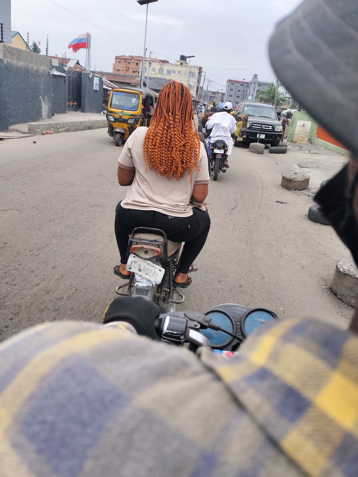 The image shows a street in Alaiyabiagba, Nigeria, taken from the perspective of someone riding a motorcycle. A woman with orange braided hair sits as a passenger on the motorcycle in front. She is wearing a beige shirt and black pants. The motorcycle's license plate is partially visible and reads "AKO 801QL." In the background, a yellow tricycle vehicle, other motorcycles, and a black SUV are visible on the street. The street is paved but shows signs of wear and tear. Buildings and a flag pole can be seen in the distance, and there are concrete barriers on the side of the road. The foreground shows parts of the motorcycle's handlebars and the rider's gloved hand and plaid shirt. The sky is overcast.
