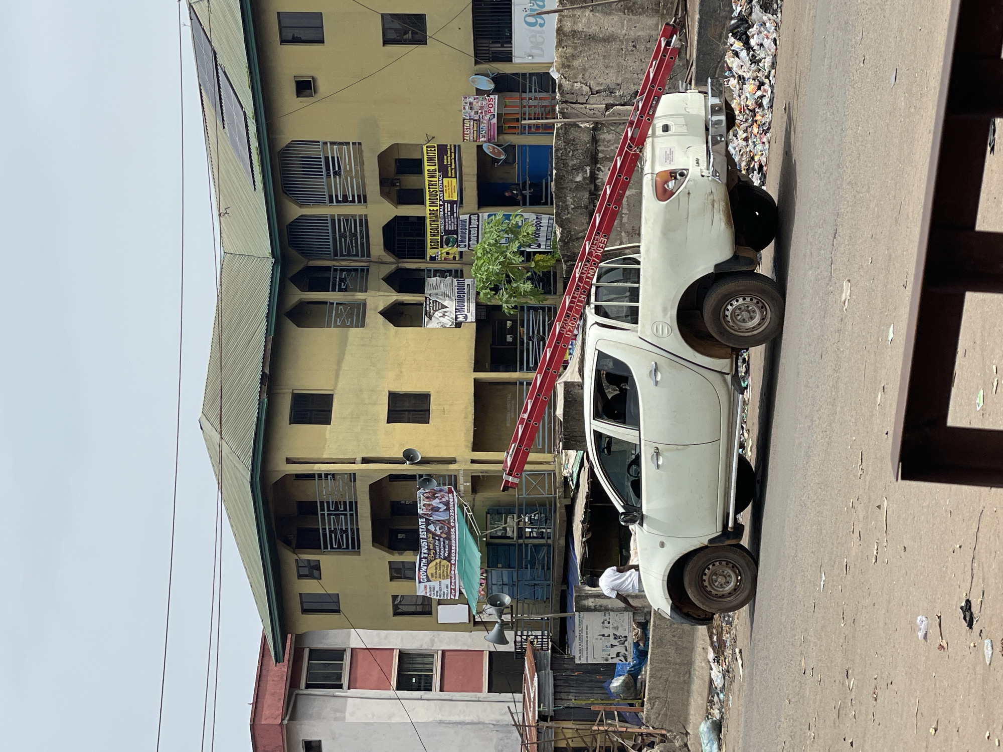 Captured in Nkpor, Nigeria, the image showcases a street scene juxtaposed with a multi-story yellow building. A white pickup truck is parked alongside the road, a red ladder resting against it. The building is adorned with various signs and advertisements, some of which read "GROWTH TRUST ESTATE", "Moniepoint", and "HEALTHCARE INDUSTRY NIG. LIMITED". A man in a white shirt is visible near the truck. Accumulations of trash line the side of the road and the buildings' curb, indicative of local waste management challenges. Overhead cables and wires are visible against a somewhat overcast sky.