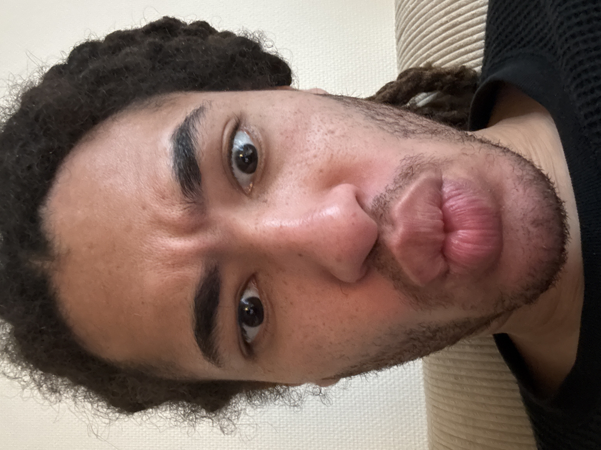 A close-up, slightly canted, selfie of a person with brown skin, dark brown eyes, and dark brown hair with locs. The person is looking directly at the camera with slightly furrowed brows and pursed lips. They have a trimmed beard. The background is out of focus; on the left, a white or cream-colored wall is visible, and on the right, there is a beige ribbed surface. The person is wearing a black, knit garment. The location is not discernible from the image.