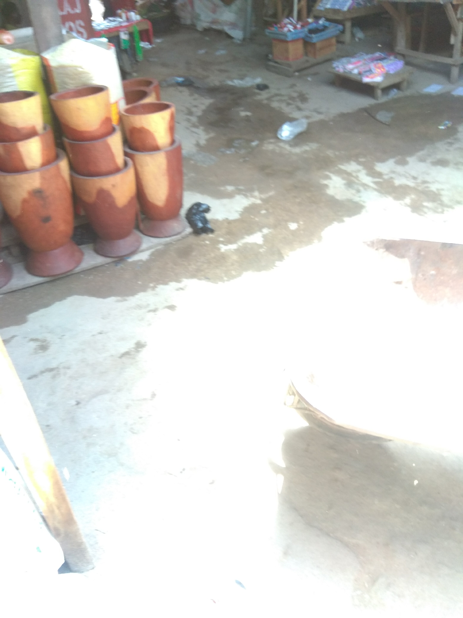 The image depicts an outdoor scene, likely a marketplace, in Jalingo, Nigeria. In the foreground, a collection of cylindrical wooden objects, resembling mortars, are stacked. These objects are a reddish-brown color with lighter, unfinished wood at the top. To the left, a portion of a large, white woven sack is visible. A small, dark object, possibly an animal or debris, rests on the wet ground to the right of the mortars. The ground is damp and appears to be a mix of concrete and dirt, with puddles of water and scattered debris. In the background, a market stall is visible with shelves displaying various items, including what appear to be packaged goods and colorful objects. The overall lighting suggests it may be daytime, with a strong glare in the upper right portion of the image obscuring some details.