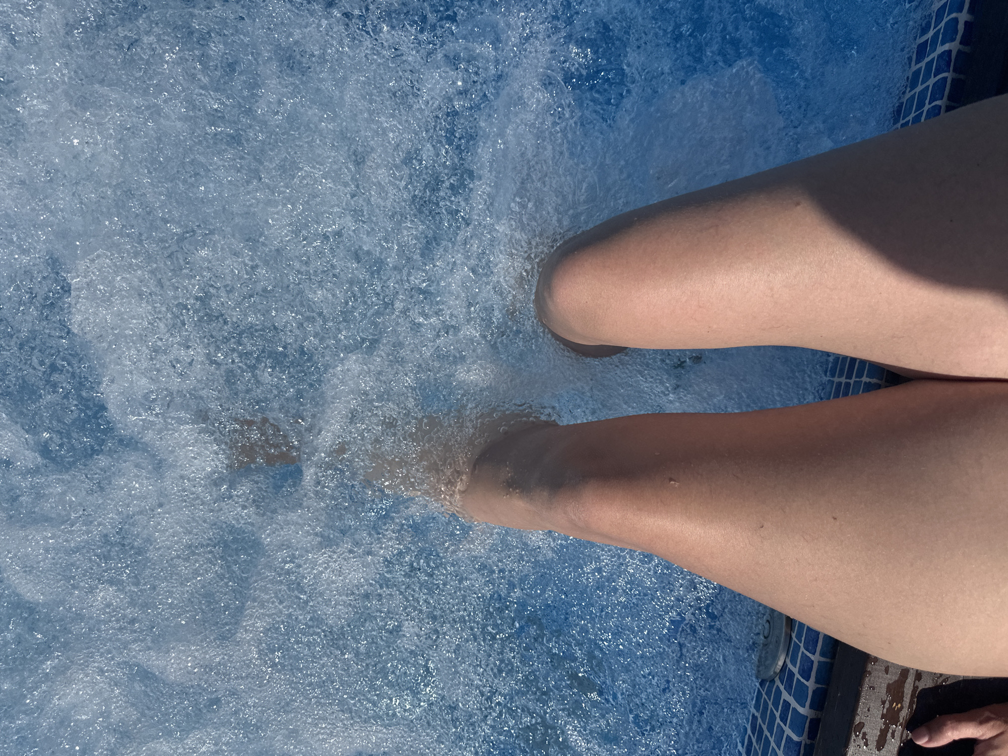 The image shows a person's legs submerged in a bubbling blue hot tub. The water is filled with numerous small bubbles, indicating that the jets are active. The person's skin appears slightly tanned and smooth. The edge of the hot tub is visible, featuring blue tiled edging and a wooden deck. A part of a foot with painted toenails is visible in the lower left corner of the image, suggesting the person is in the hot tub. The environment is likely an outdoor setting, given the bright lighting and the hint of a shadow on the person's right leg. The location is stated to be in the city of Danilovka (Altai), Russia, implying the hot tub is situated within or near this area.