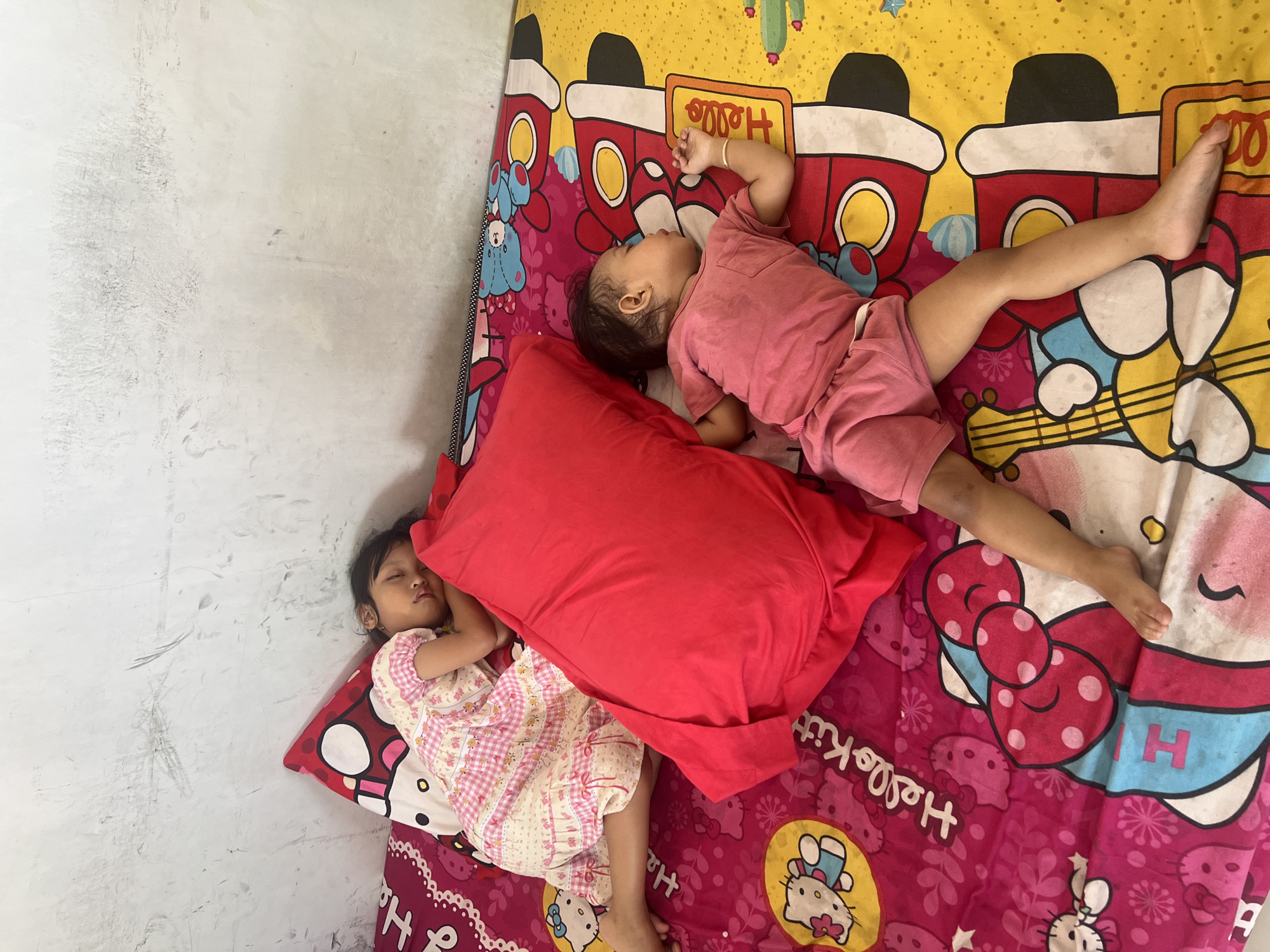 In a bedroom in Kecamatan Pati, Indonesia, two young children are asleep on a bed. The child on the left, a girl, wears a pink and white checkered dress and is lying on her side, propped up by a red pillow and a Hello Kitty pillow. The child on the right, a boy, wears a pink t-shirt and shorts and is lying on his back. Both children are on a bed with a colorful bedspread featuring cartoon characters and musical instruments. The wall behind the bed is a light gray, textured surface.
