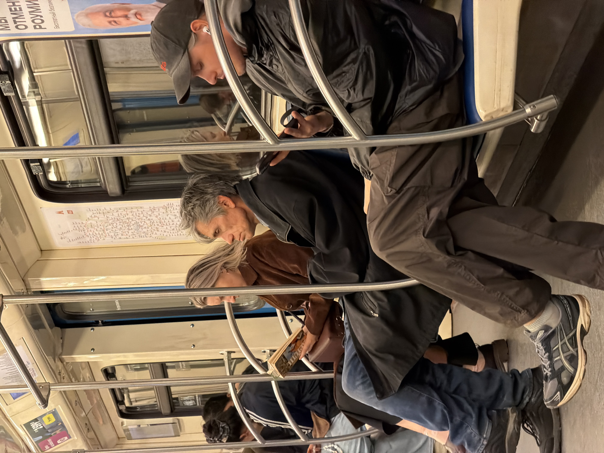 The image shows the interior of a Moscow Metro train car in Russia. Several people are seated, with some focused on their phones or reading. The passengers are dressed in casual clothing, suggesting an ordinary commute. The environment includes standard subway car elements, such as windows with advertisements, handrails, and seat backs. The train car's interior appears clean and well-maintained.
