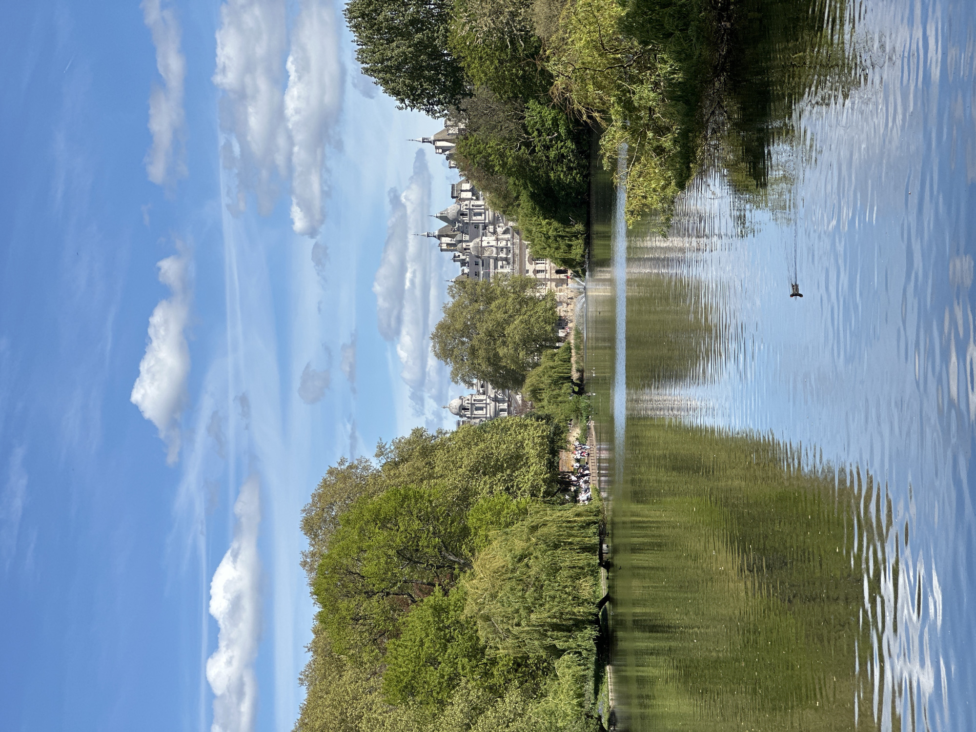 The image depicts a park scene with a body of water, likely a lake or large pond, reflecting the surrounding trees and sky. The water's surface is rippled, indicating a slight breeze. On the opposite bank, a large building with multiple towers and a pale facade is visible through the trees. Along the edge of the water, a group of people is gathered, possibly picnicking or relaxing. The sky is blue with scattered white clouds. A bird is seen swimming in the water. The location is likely St. James's Park in the City of London, United Kingdom, and the building is likely the Foreign, Commonwealth & Development Office.