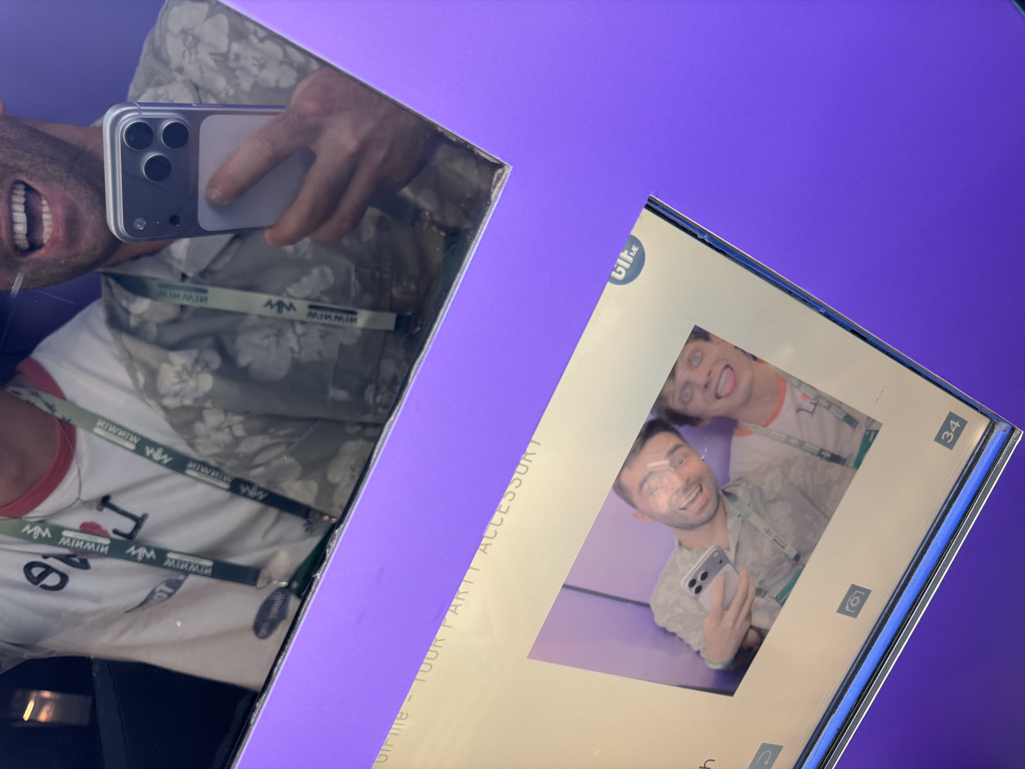 The image shows a mirror reflection of two individuals, likely in an indoor setting with purple walls. The person in the foreground is taking a selfie with a smartphone, their mouth open in a wide smile. They are wearing a light-colored short-sleeved shirt with a floral pattern and a lanyard around their neck. A portion of another person's face is visible to the right, also smiling broadly and sticking out their tongue.
Below the mirror reflection, there is a framed print or screen displaying a photo booth-style image of the same two individuals. In this photo, the person on the left is holding up a smartphone and smiling, while the person on the right is making a similar facial expression with their tongue out. The print also includes what appears to be a logo or text that reads "GIRLME - YOUR PARTY ACCESSORY" and a number "34". There are also small icons that suggest a digital display, possibly from a photo booth.
The environment suggests a commercial or event space, possibly a photo booth at a party or event. There is no indication of the city of Tbilisi, Georgia, in the image.
