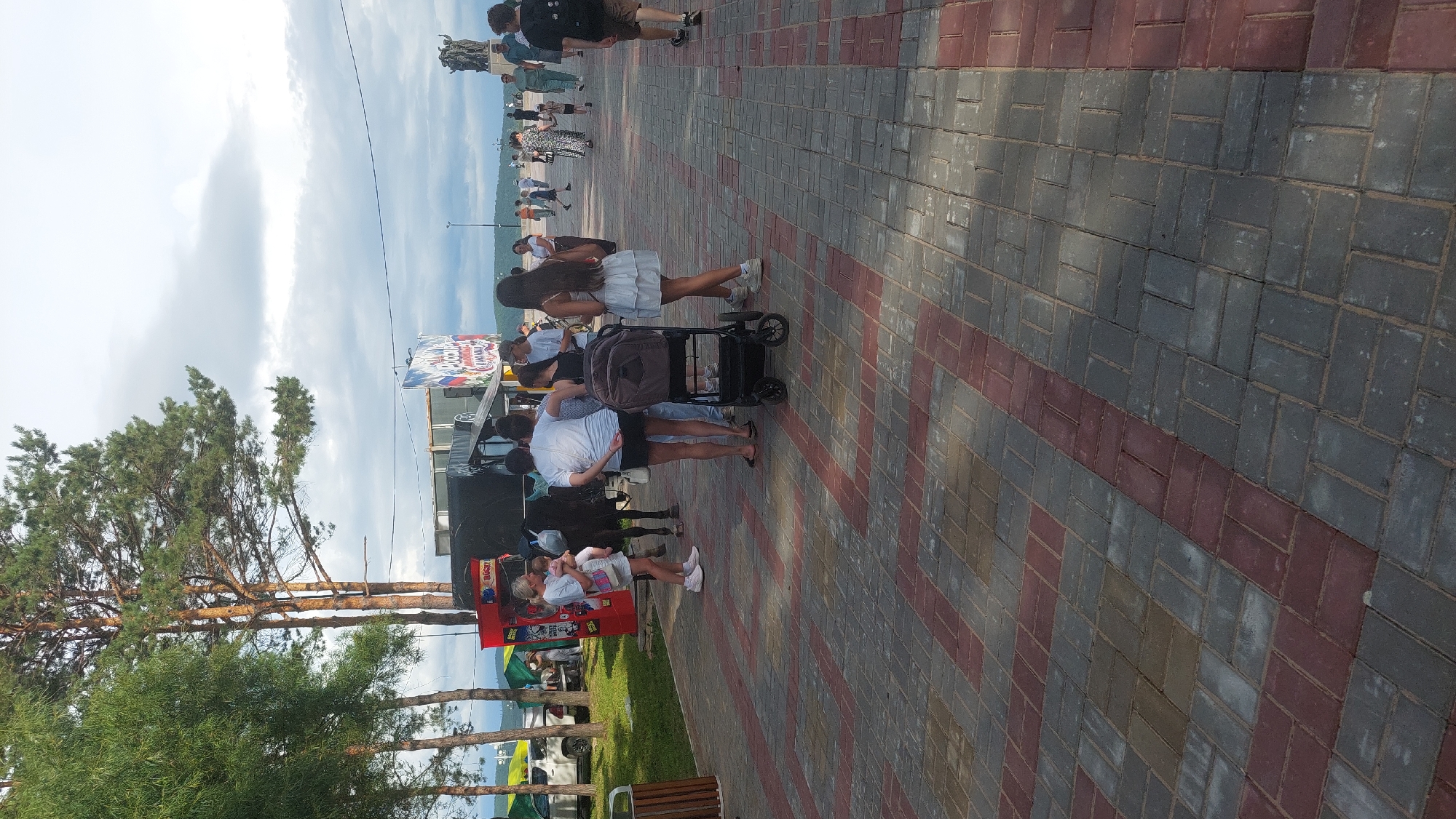 A full-body, eye-level, vertical shot depicts a paved walkway with a red and gray brick pattern, extending into the distance. The scene is populated by several individuals and some objects. In the foreground, a stroller with a dark gray frame and fabric sits unoccupied. A woman with long, dark hair, wearing a white dress, stands near the stroller. To her left, a group of people are gathered around what appears to be a black horse, possibly for a ride or interaction. A woman in a white shirt and black shorts is holding a baby. Another woman in a white shirt and white shorts is also present, holding a white bag. To the far left, a red vending machine stands near a tree line. In the midground, a few people are walking along the path. Beyond them, a body of water is visible with a distant shoreline and a cloudy sky overhead. The background features a statue on the right side, a large banner with Russian text and flags on a structure, and trees lining the left side of the image. The overall atmosphere suggests a public gathering or event in an outdoor urban setting, possibly a park or embankment, in Komsomolsk-on-Amur, Russia, given the Russian text on the banner.