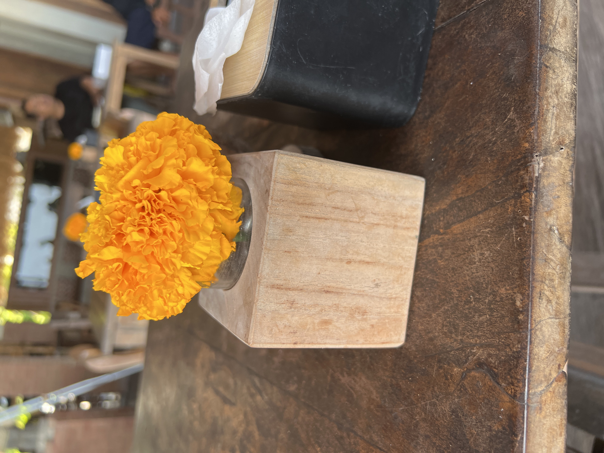A bright orange marigold is in a small glass vase, which is placed in a square, light-colored wooden holder. This holder sits on a dark brown, weathered wooden table. To the right of the marigold is a black tissue box with a wooden top, from which a white tissue protrudes. In the background, out of focus, are people sitting at tables in what appears to be an outdoor cafe or restaurant. The setting is likely a cafe in North Kuta, Indonesia, suggested by the marigold, commonly used in offerings and decor in Bali.