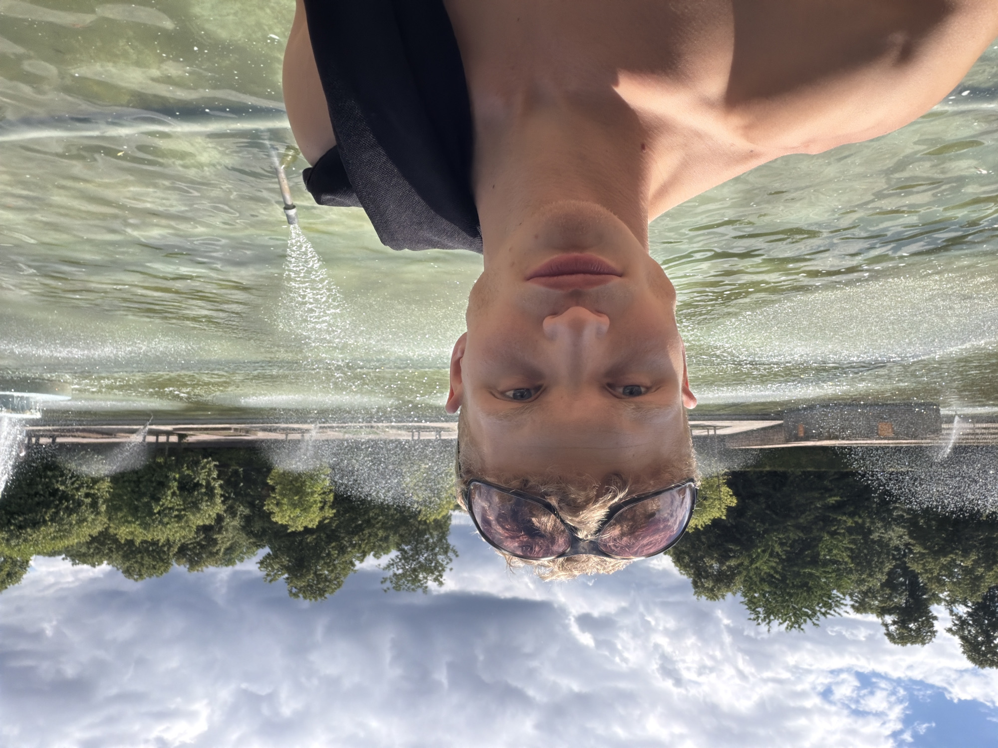 The image is an upside-down, close-up selfie of a person with fair skin and blonde hair, wearing sunglasses. They are in what appears to be a park or garden setting, with a body of water and fountains visible in the background. The water is reflecting the sky, which is partly cloudy. Trees and some architectural elements are also visible in the distance. The person's face is centered in the frame, and they are looking directly at the camera. The lighting suggests it is daytime, and the overall mood seems casual. No text is visible in the image.