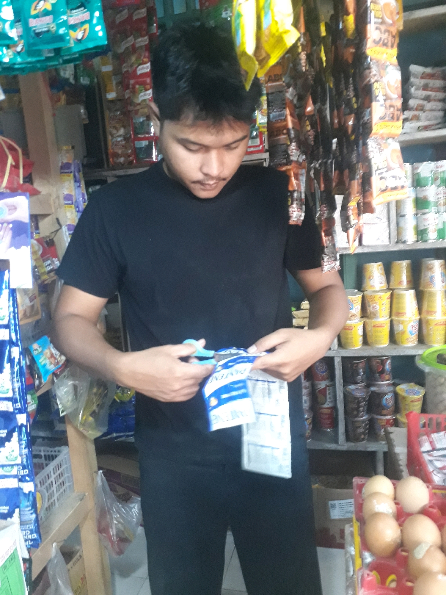 A young man wearing a black t-shirt and black pants is standing in what appears to be a small convenience store or warung in Pangkalpinang, Indonesia. He is holding a pair of light blue scissors and is in the process of cutting open a white and blue packet. The store is filled with shelves stacked with various packaged goods, including food items like instant noodles in cups and bags, as well as what appear to be snacks and drinks. The lighting is somewhat dim, suggesting an indoor environment. In the foreground, to the right, a red plastic basket holds several eggs. The overall impression is that of a busy, local retail setting.