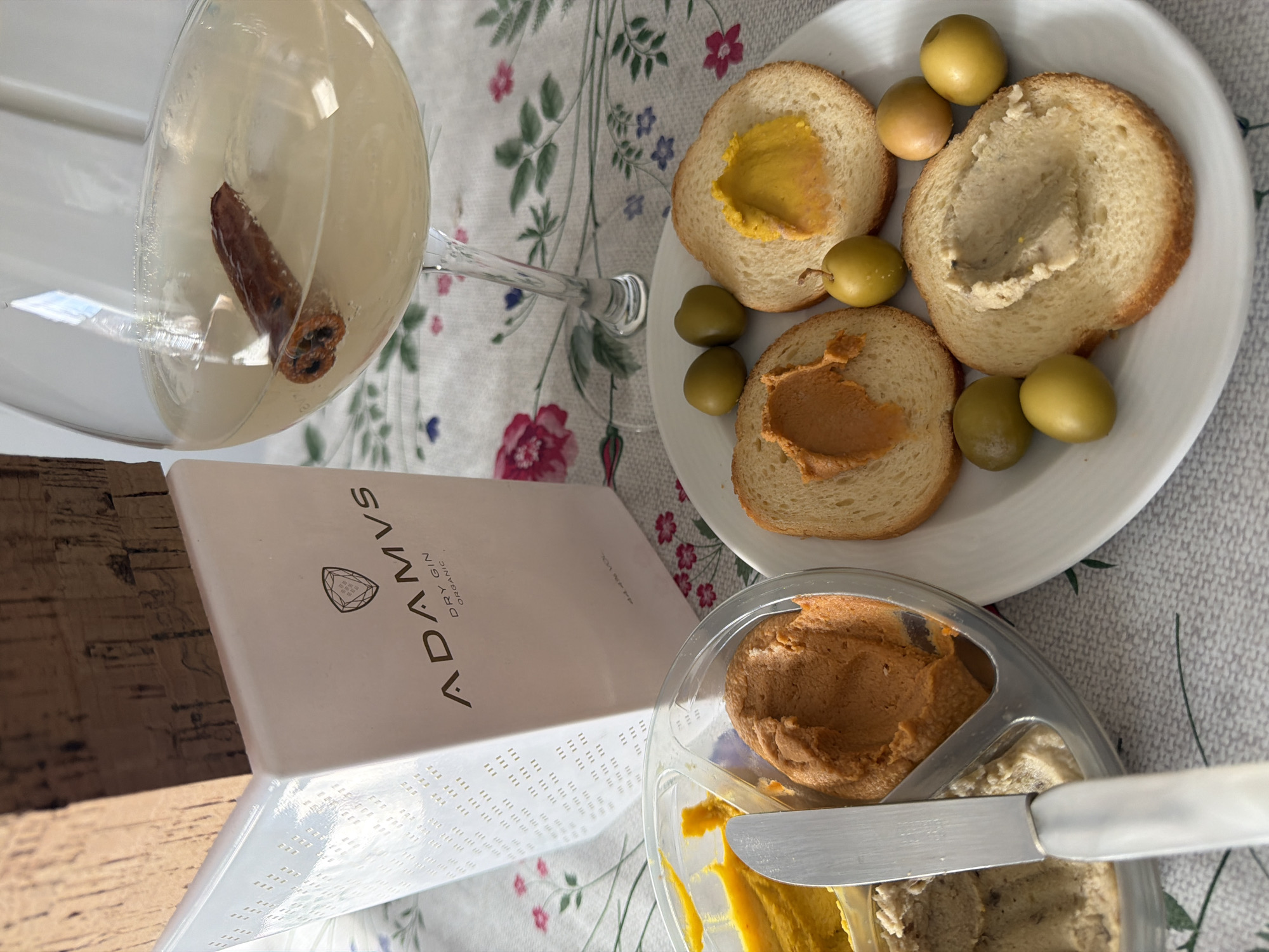 An overhead, slightly angled photograph depicts a tabletop spread, likely an appetizer or snack, located in Esch-sur-Alzette, Luxembourg.
In the foreground, a transparent plastic multi-compartment container holds three distinct spreads: a vibrant yellow, an orange, and a light beige. A silver spreading knife with a white handle rests across the yellow and orange spreads. To the right, a white, slightly oval plate features three slices of toasted bread, each topped with one of the aforementioned spreads. Six whole, green olives are distributed around the bread on the plate.
In the midground, a tall, white, rectangular bottle with a large, textured cork stopper is visible. The bottle is labeled "ADAMVS DRY GIN ORGANIC" with a geometric logo positioned above the text. Its left side features a grid of small, square perforations. To the right of the bottle, a clear stemmed wine glass contains a pale yellow liquid and a single cinnamon stick submerged within it.
All items are arranged on a light-colored tablecloth adorned with a floral pattern consisting of pink and blue flowers and green leaves, set against a light grey or beige textured background. No individuals are visible in the scene.
