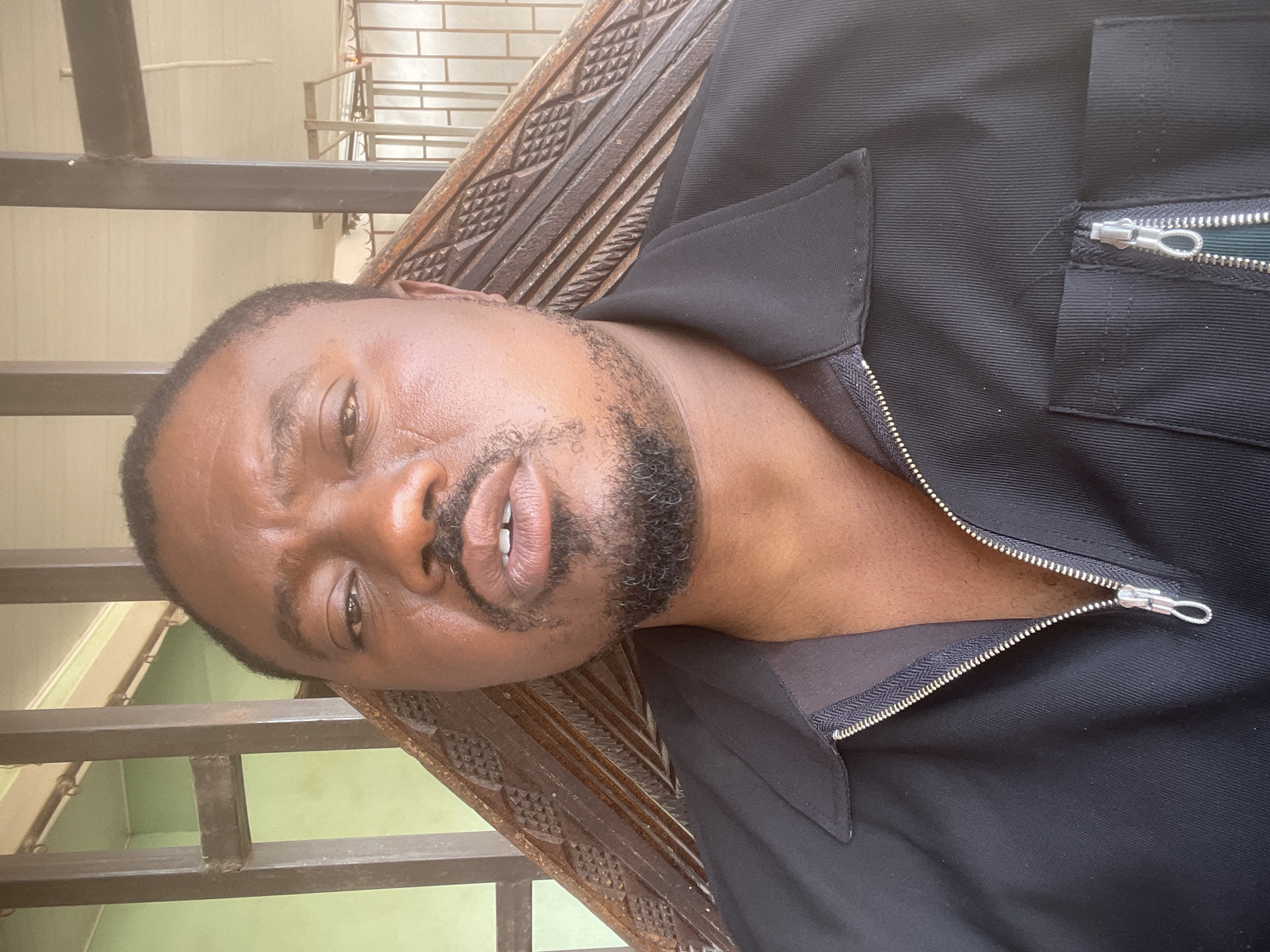 A man with dark skin, short black hair, and a neatly trimmed black beard is looking directly at the camera. He is wearing a black zip-up shirt with a collar. He appears to be seated on a wooden bench with decorative carvings. A metal railing is visible behind him, suggesting an outdoor setting. The image was taken in Barakin Akawo, Nigeria.