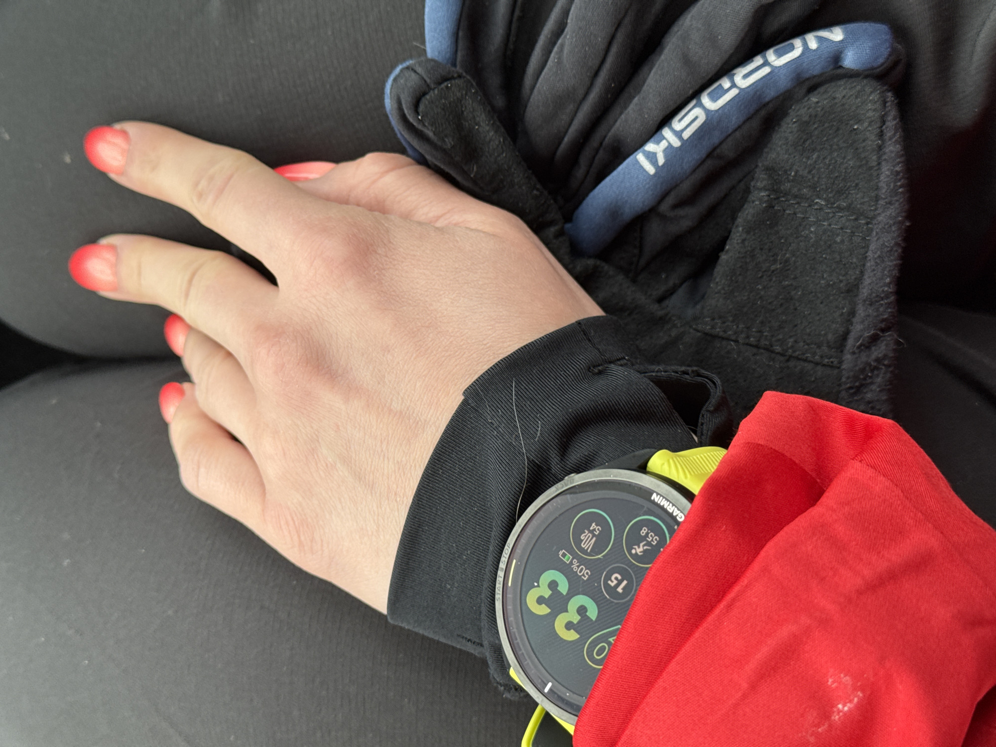 A person's left hand rests on their leg. The hand has bright red-tipped nails. A black glove with blue lettering that reads "NORDSKI" is on top of the hand. A black long-sleeved shirt covers the wrist, and a Garmin watch with a yellow band is visible on the wrist, partially obscured by a red shirt. The watch face displays "33" in large green numbers. The leg has a dark material. The location is not specified, and there is no indication that the image was taken in Chaikovsky, Russia.