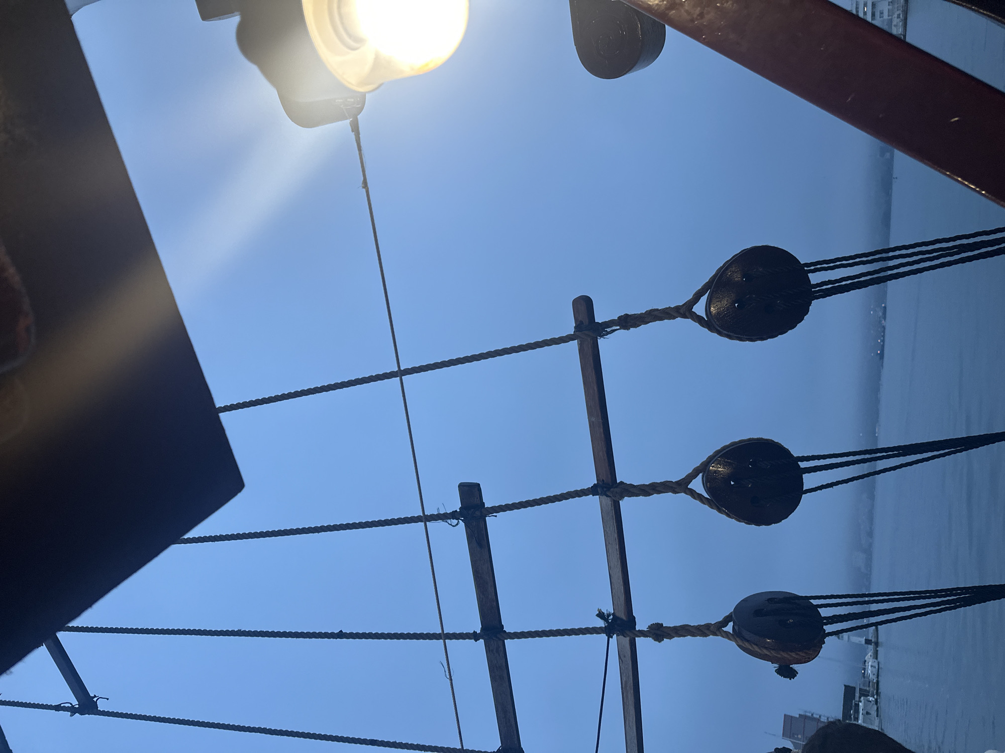 The image is an upward-facing view of the rigging on a sailing ship, including ropes, wooden spars, and pulleys. A bright light fixture is visible at the top left. Beyond the ship’s structure, a body of water is visible, along with a blurred urban landscape in the distance. Given the subject matter, the location may be Varna, Bulgaria, given the presence of a maritime museum and harbor. The photo seems to have been taken at dusk or during an overcast day.