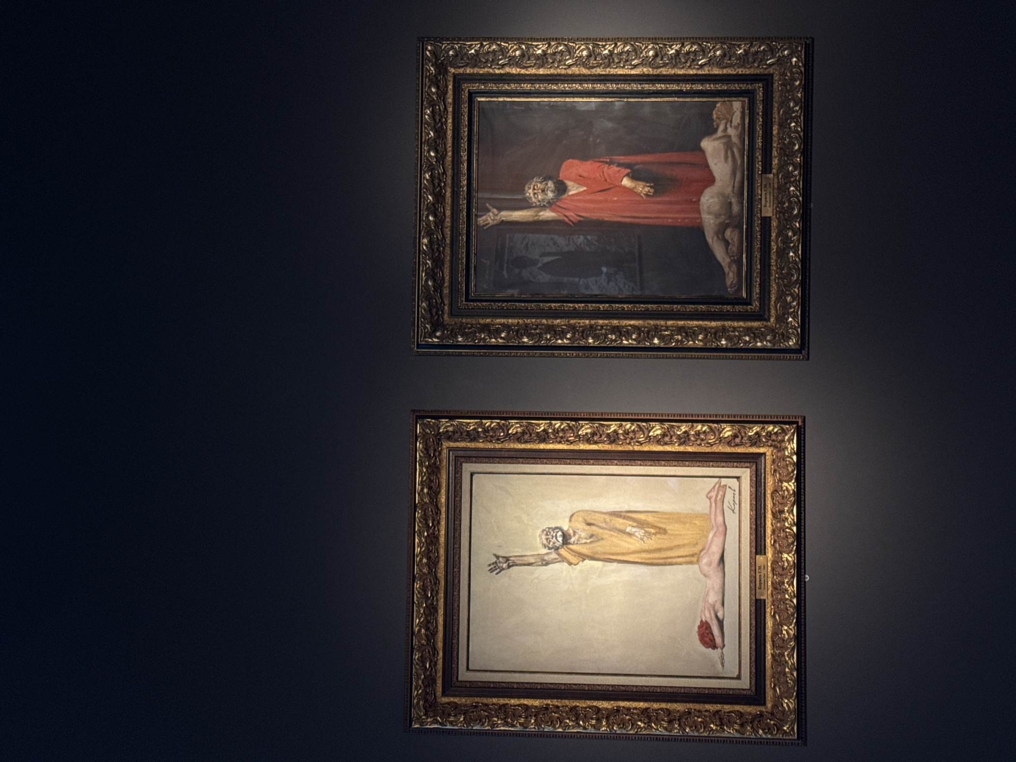 Two framed paintings hang on a dark gray wall. The top painting depicts a man in a red robe with his arm outstretched, next to a nude figure. The bottom painting shows a similar scene with a man in a yellow robe, also with an outstretched arm, adjacent to a nude figure. Both paintings are in ornate, gold-colored frames. The location is likely a museum or gallery in Moscow, Russia, based on the artwork style.