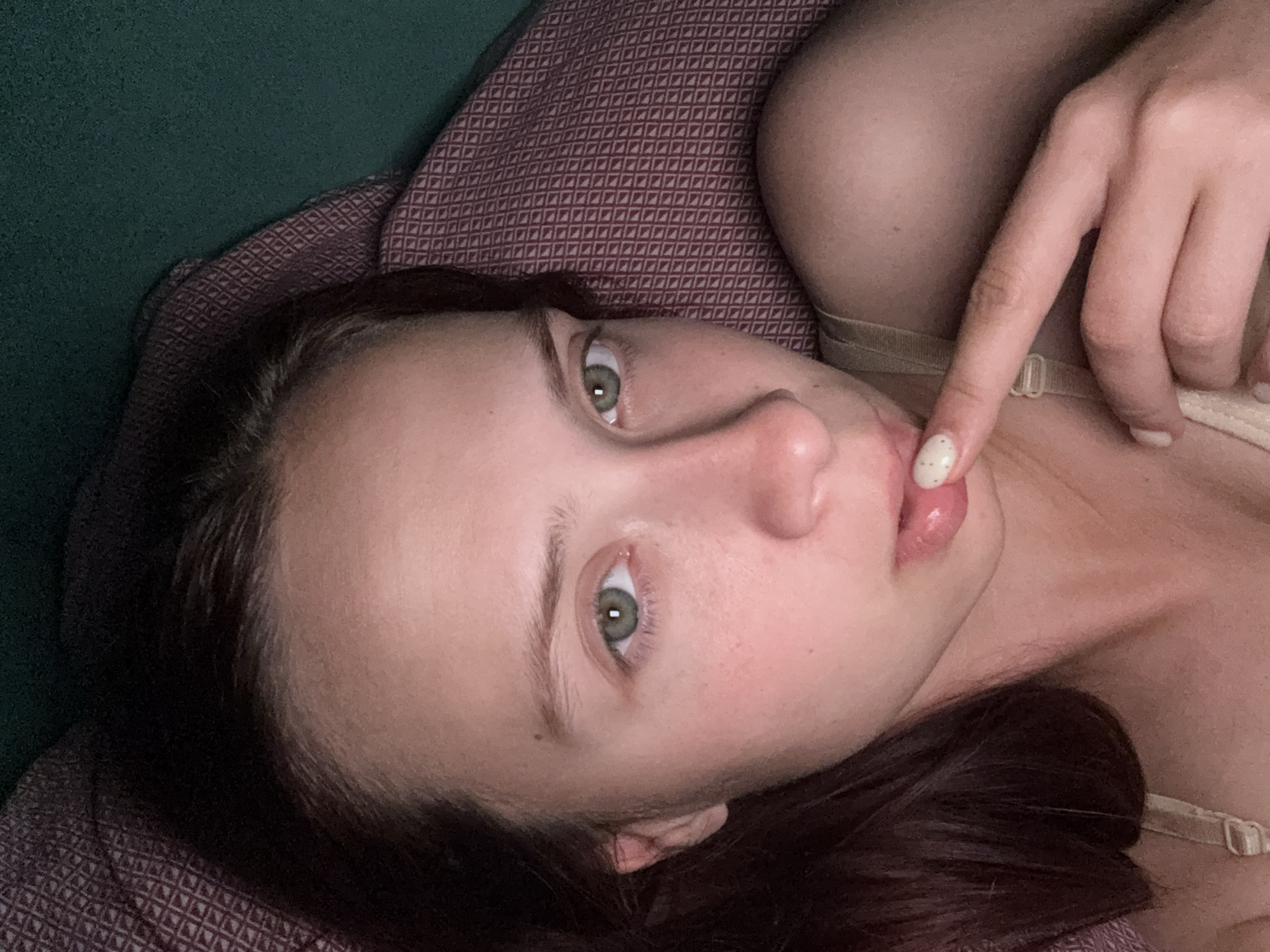 The image is a close-up, first-person perspective photograph of a young woman with long, dark hair and green eyes. She is lying down, with her head resting on a pillow that has a dark red geometric pattern. A portion of a green surface is visible behind the pillow. The woman is wearing a light-colored tank top or bra. Her right hand is raised, with her index finger touching her lips. Her fingernails are painted with a light color and small dots. The lighting suggests an indoor setting, possibly a bedroom. There is no discernible indication of the specific location within the city of Прикамский, Russia.