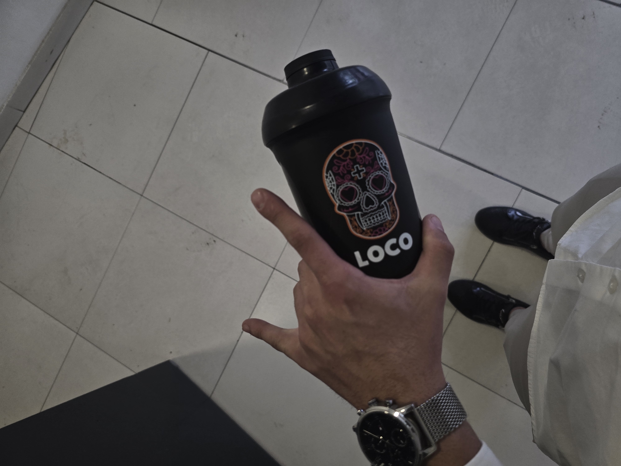 This overhead photograph captures a person's hand holding a black shaker bottle against a light-colored tiled floor. The shaker bottle features a colorful sugar skull design with intricate pink, orange, and white patterns, and the word "LOCO" printed below it in white capital letters. The hand displays a gesture with the pinky and index fingers extended, resembling a "shaka" or "devil horns" sign. A silver-toned watch with a mesh band and a dark, multi-dial face is visible on the person's left wrist. In the lower right of the frame, part of a white garment, possibly a shirt or trousers, is seen along with two black, shiny shoes. A dark, angular surface occupies the bottom left corner of the image. The setting, characterized by the tiled floor and artificial lighting, suggests an indoor environment within Sofia, Bulgaria, such as a corridor, hall, or commercial space. No specific activity beyond holding the bottle and standing is clearly depicted.