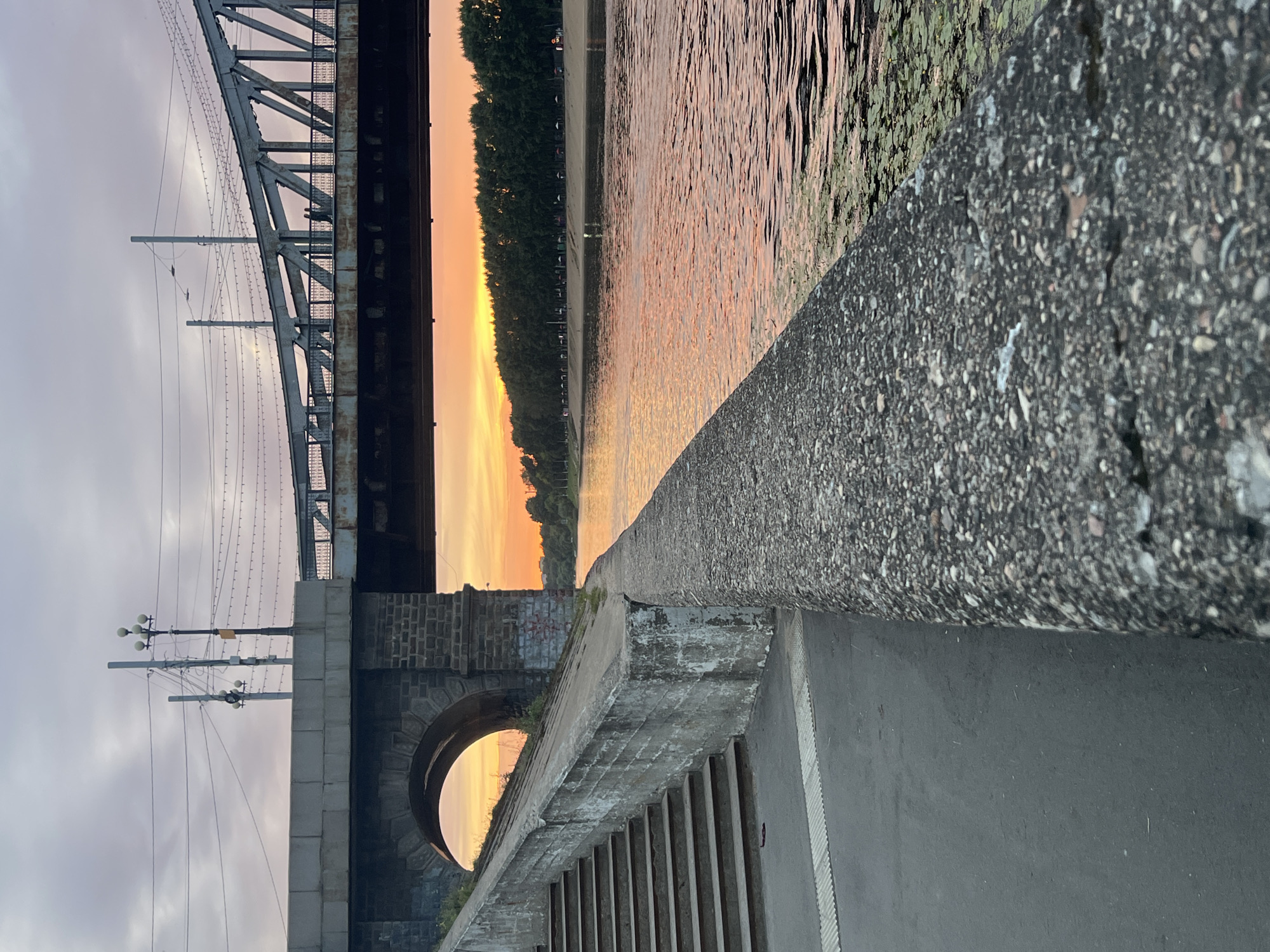 The image depicts a sunset over the Volga River in Tver, Russia, viewed from the embankment. A stone embankment wall, weathered and textured, occupies the foreground, leading the eye towards the river. To the left, a concrete staircase ascends along the side of a stone bridge abutment, which features an arched opening. The bridge itself, a metal truss structure, spans the river, with indistinct figures visible walking on its walkway. The sky is filled with soft, muted colors of dusk, with clouds streaking across the horizon. The river surface reflects the warm orange and pink hues of the sunset, with patches of green lily pads floating on the water. A line of trees borders the opposite bank, with a few small structures or tents visible along the shore. Electrical wires and streetlights are visible on and around the bridge, suggesting an urban environment. The scene is serene and captures the tranquil beauty of a sunset by the river.