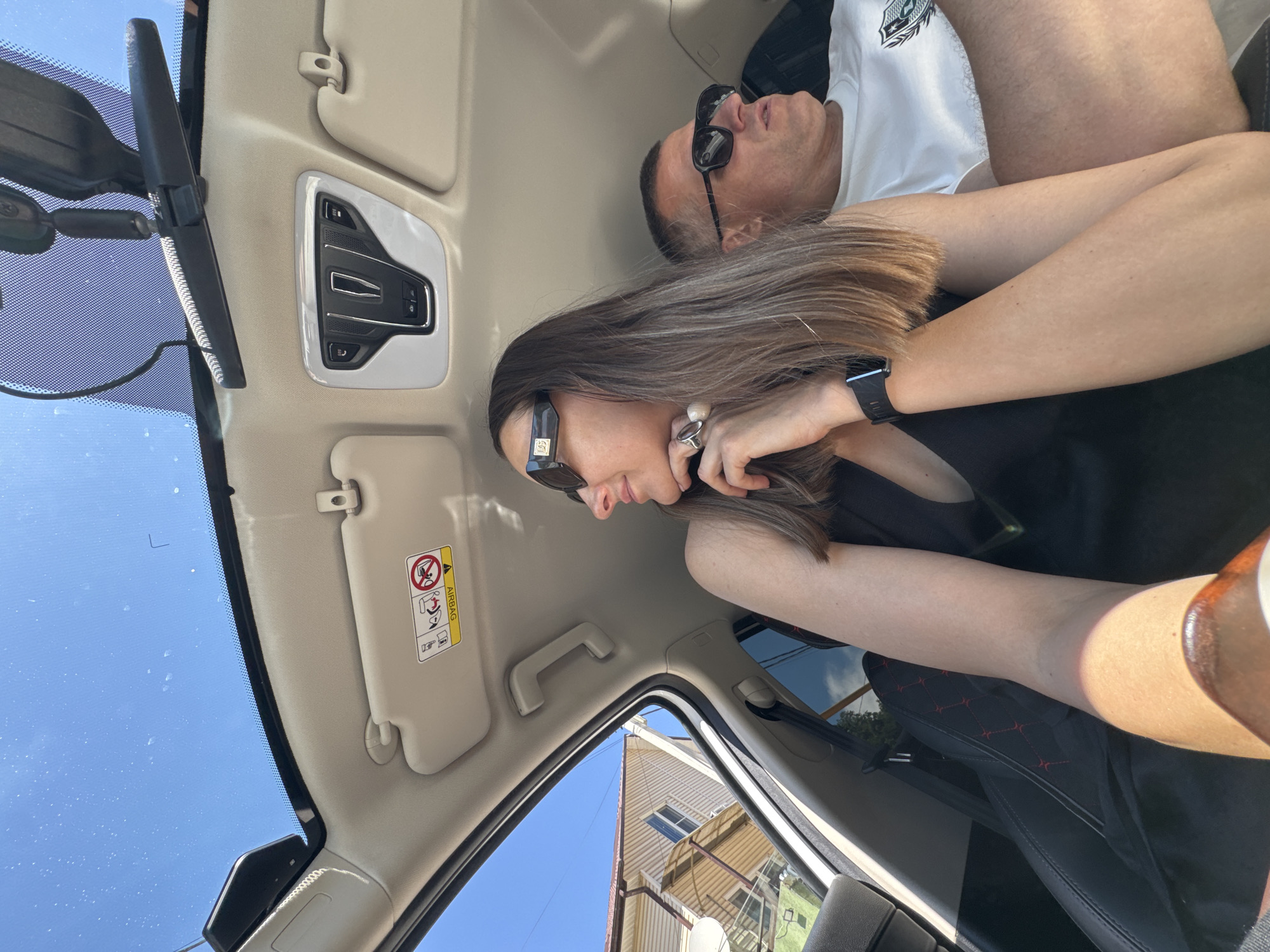An image captures the interior of a vehicle during daylight, presumed to be in Sochi, Russia. The perspective is from within the car, looking towards the front and left.
In the foreground, a woman is seated as a passenger, looking to her left out the vehicle's window. She has long, light brown hair with lighter highlights, and is wearing a black sleeveless garment. Dark sunglasses with a "D&G" logo on the temples obscure her eyes. A dark smartwatch is visible on her left wrist, and a large, light-colored ring is on her right ring finger, which is resting against her chin.
To her right and slightly behind, a man is also seated as a passenger, looking to his right. He is wearing a white short-sleeved t-shirt with a dark emblem on the left chest and dark sunglasses. His left arm is visible next to the woman's right arm.
The car's interior features a light-colored headliner and sun visors, one of which displays a yellow "AIRBAG" warning label. An overhead console unit with multiple buttons, including "SOS," is centrally located. A rearview mirror hangs from the windshield. A large glass sunroof or panoramic roof is visible overhead, showing a clear, bright blue sky with scattered small marks (possibly dust or water spots) on its surface. Strong sunlight enters the vehicle, creating defined shadows.
Outside the left side of the vehicle, a multi-story residential building with light-colored horizontal siding and a white satellite dish is visible under the clear blue sky. Green foliage is also discernible in the distance through the side windows. A black car seat with red diamond-pattern stitching is partially visible on the lower left of the frame.
