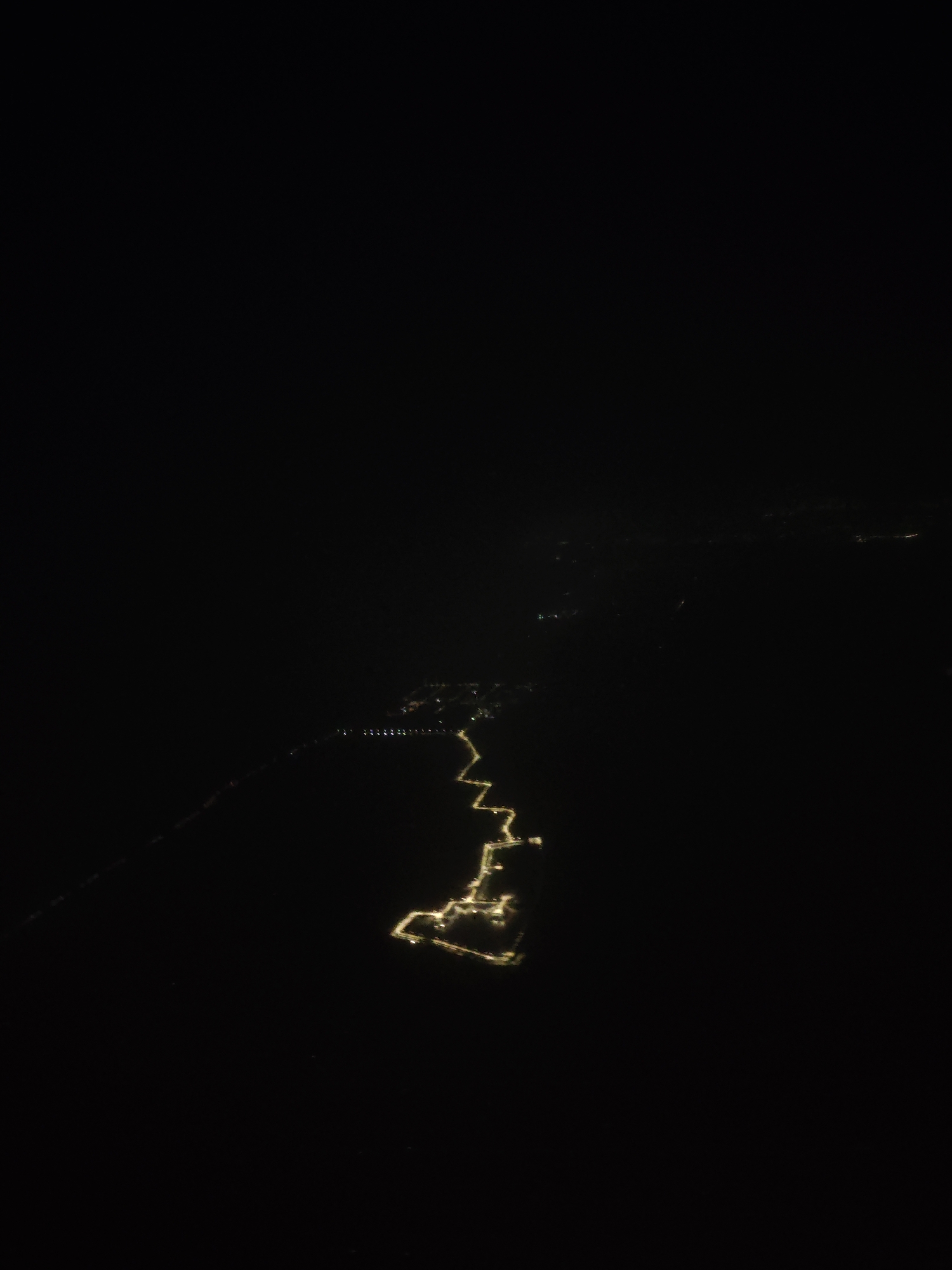 The image shows a night view of Novosibirsk, Russia, from a high vantage point, possibly from an aircraft or a tall structure. The city is visible through a trail of yellow-orange street lights tracing its roadways and urban areas, illuminated against the surrounding darkness. There are a few sparsely lit neighborhoods and isolated structures. In the far distance there is a faint greenish glow which seems to be from some sort of city light or a park.