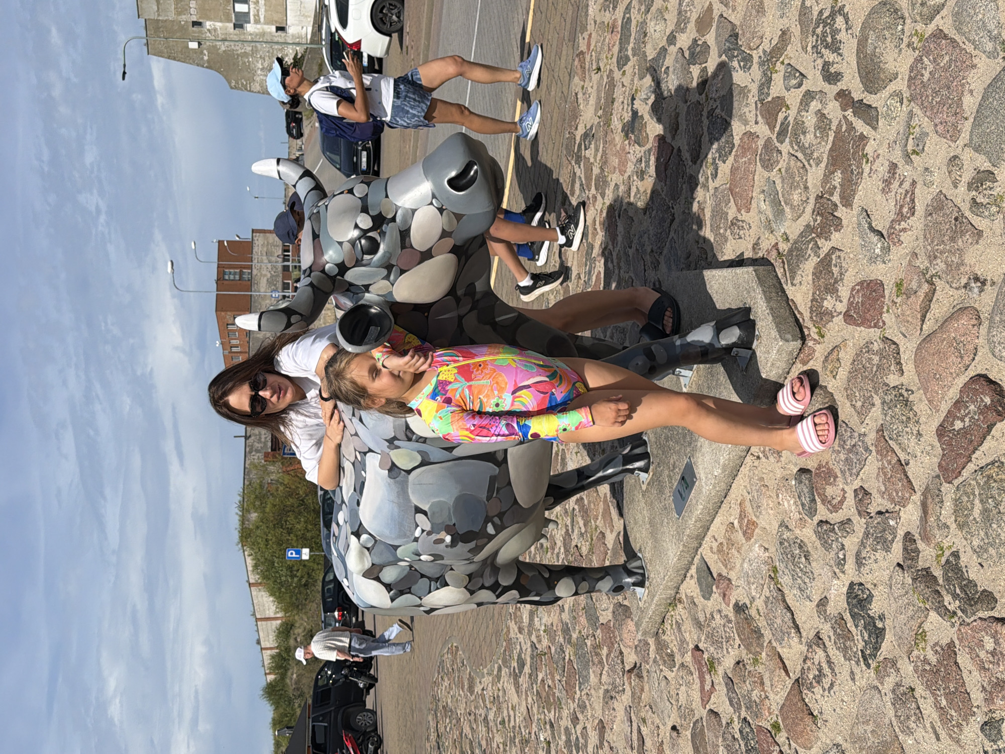 In Ventspils, Latvia, a young girl wearing a colorful swimsuit and pink slides stands next to a sculpture of a cow. The cow sculpture is decorated with a mosaic of grey and white stones. A woman, presumably the girl's mother, is leaning on the cow sculpture behind her. The scene is set outdoors on a cobblestone path with sand. In the background, there are parked cars, buildings, and streetlights under a partially cloudy sky.