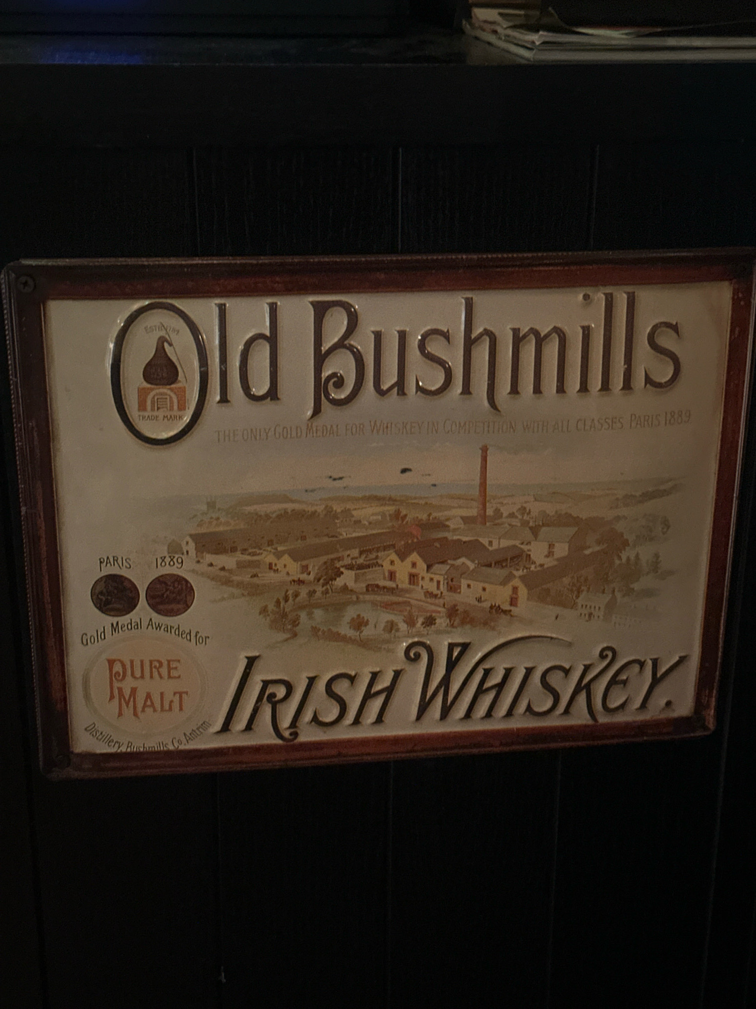 The image shows a close-up of a rectangular metal sign for "Old Bushmills Irish Whiskey." The sign features a depiction of a distillery with a tall chimney. The text on the sign indicates "Established 1869," "THE ONLY GOLD MEDAL FOR WHISKEY IN COMPETITION WITH ALL CLASSES PARIS 1889," and "Gold Medal Awarded for PURE MALT." There are also circular emblems showing what appear to be medals. The sign is framed with a dark red border and mounted on a dark, textured background, possibly a wooden cabinet or wall. The lighting is dim, suggesting an indoor setting. There are no people visible in the image. The sign is in good condition, with some slight wear visible. The location is stated as the city of Belgrade, Serbia.