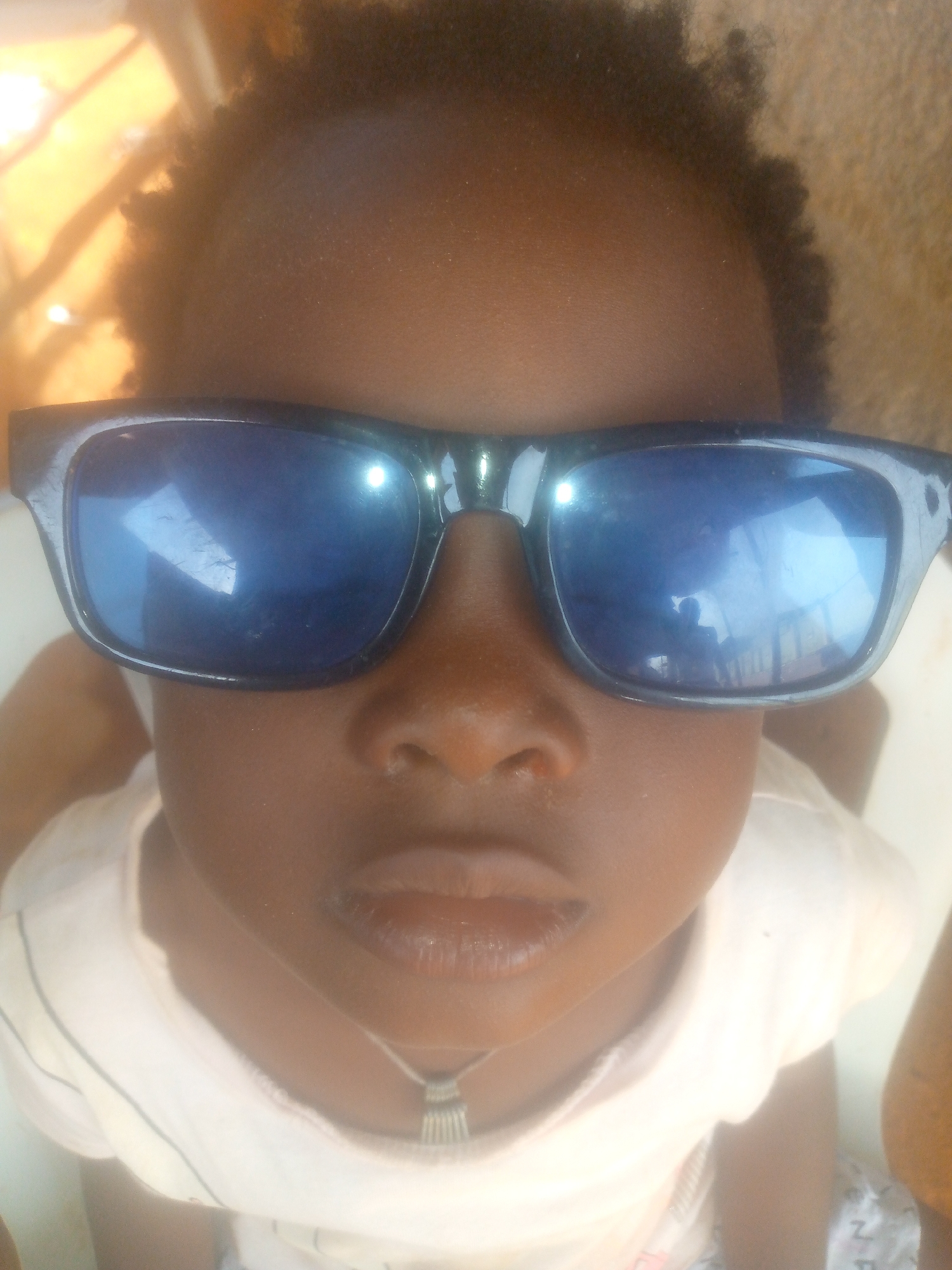 A child with dark skin and short, dark curly hair is wearing oversized black sunglasses with blue-tinted lenses. The child is wearing a white t-shirt and a silver necklace with a decorative pendant. The background is blurred and indistinct. The location is Unknown, Unknown.