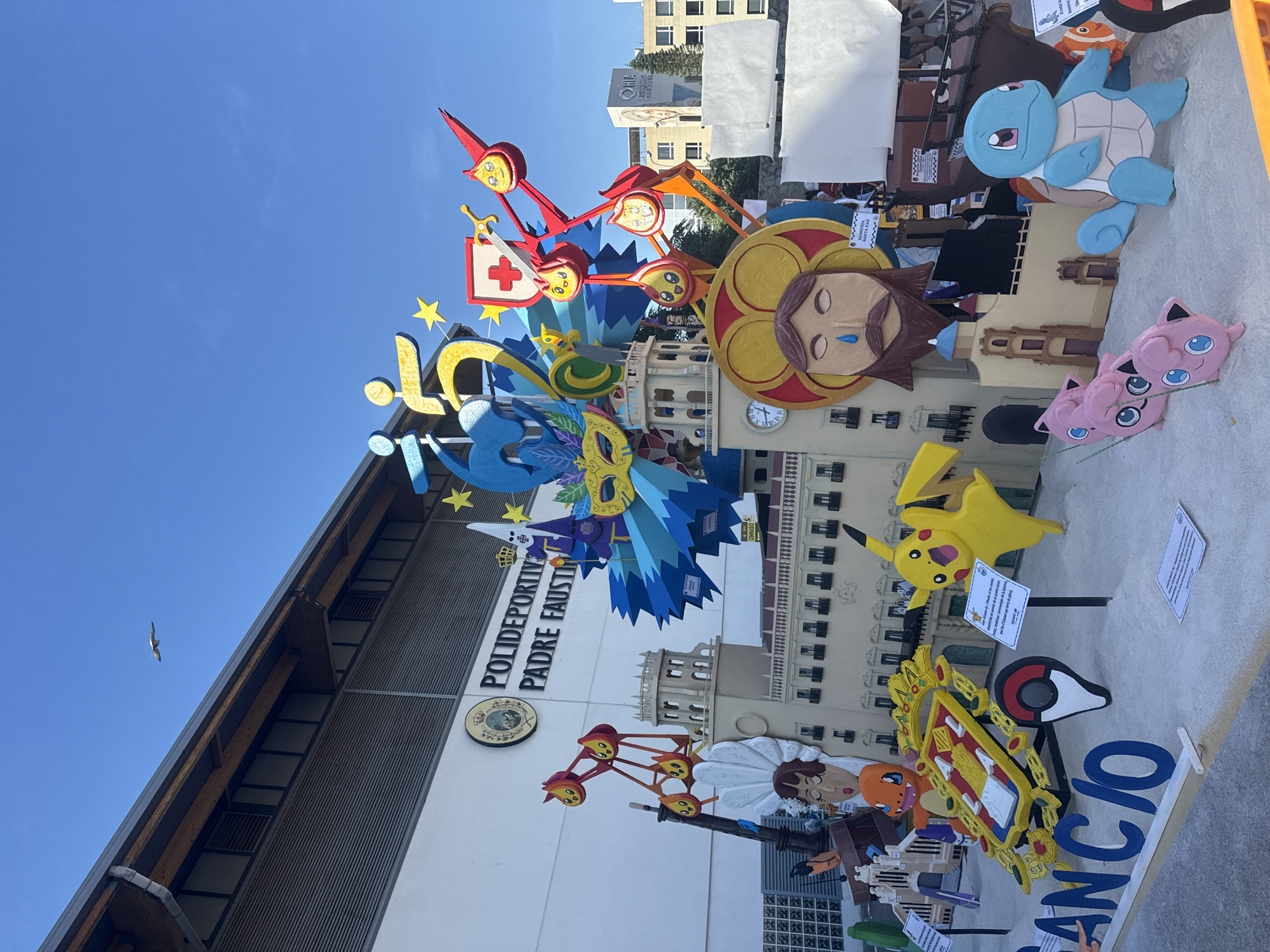 The image depicts a large, colorful outdoor sculpture in front of a building labeled "POLIDEPORTIVO PADRE FAUSTIN." The sculpture is elaborate and features various characters and structures, including what appear to be representations of Pokémon like Pikachu, Charmander, Squirtle, and Jigglypuff. There's also a depiction resembling Jesus with a tear on his cheek, a carnival mask, and figures that look like they are from a parade or festival. The sculpture is built on a sandy base, and signs with text are visible in front of some of the figures. In the background, other buildings and a clear blue sky with a bird in flight can be seen. The overall scene suggests a public event or festival, likely in a Spanish-speaking location given the text on the building.