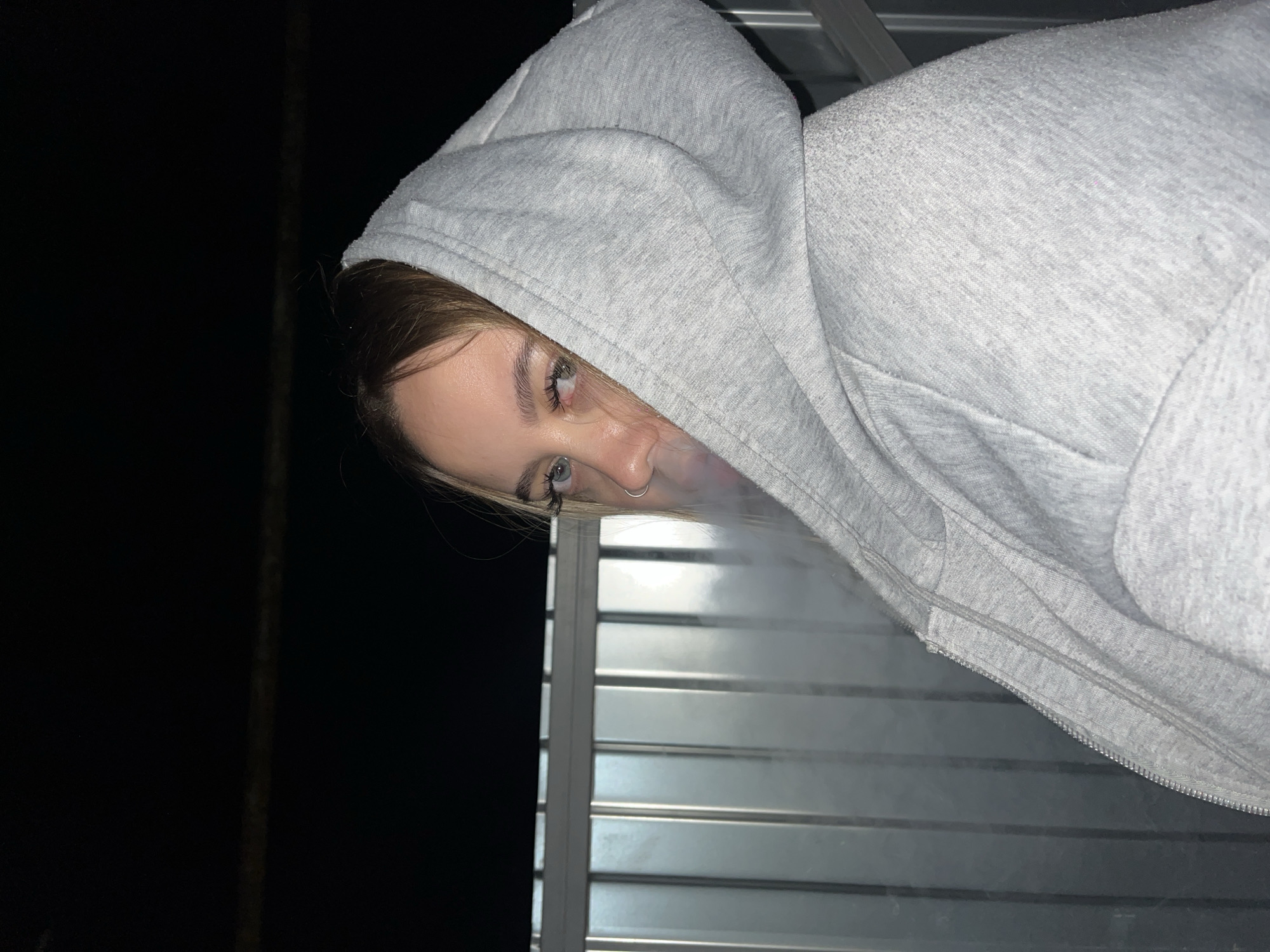 A woman wearing a grey hoodie is angled so her head and shoulders are visible in the upper right portion of the frame. She is looking toward the camera with a pierced nostril. A wisp of vapor is coming from her mouth. Behind her, a slatted silver structure is visible, and to the left, is a darkened area. There is no evidence to suggest this image was taken in Novopokrovskaya, Russia.
