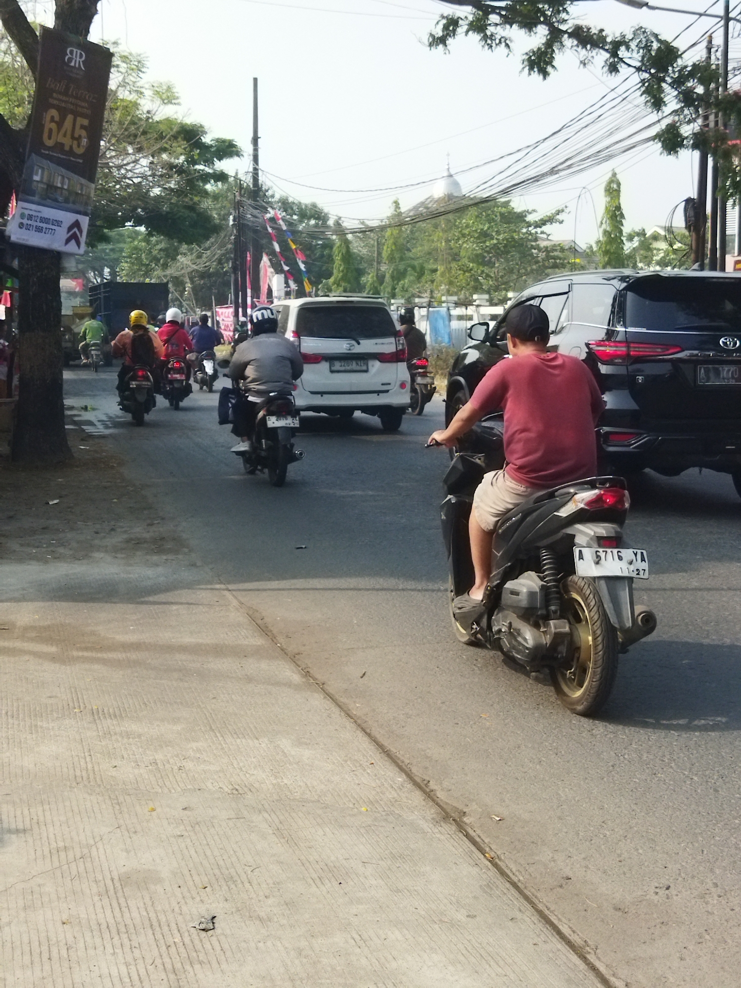 The image captures a street scene in Sepatan, Indonesia, likely on a sunny day judging by the shadows. In the foreground, a man wearing a red t-shirt and beige shorts is riding a dark-colored scooter with a gold-accented wheel. He is wearing a black baseball cap and is looking forward. The scooter has a license plate that reads "A 5716 YA" and "11.27".

Behind him, other vehicles are visible. To his left, a white SUV, possibly a Toyota Fortuner, is parked or moving slowly. Further back, a white van is also present, along with several other motorcycles carrying riders wearing helmets. Some riders are wearing yellow or red helmets. The road surface is asphalt, with a concrete sidewalk on the left side of the image.

The environment includes trees lining the street, some with lush green foliage. Overhead, there are numerous electrical wires and poles. A tall advertising banner is attached to a tree on the left side of the frame, displaying text and graphics, including the number "645". Flags or banners are strung across the street in the background, adding to the urban atmosphere. The overall impression is that of a busy street with typical motorized traffic in a Southeast Asian urban setting.