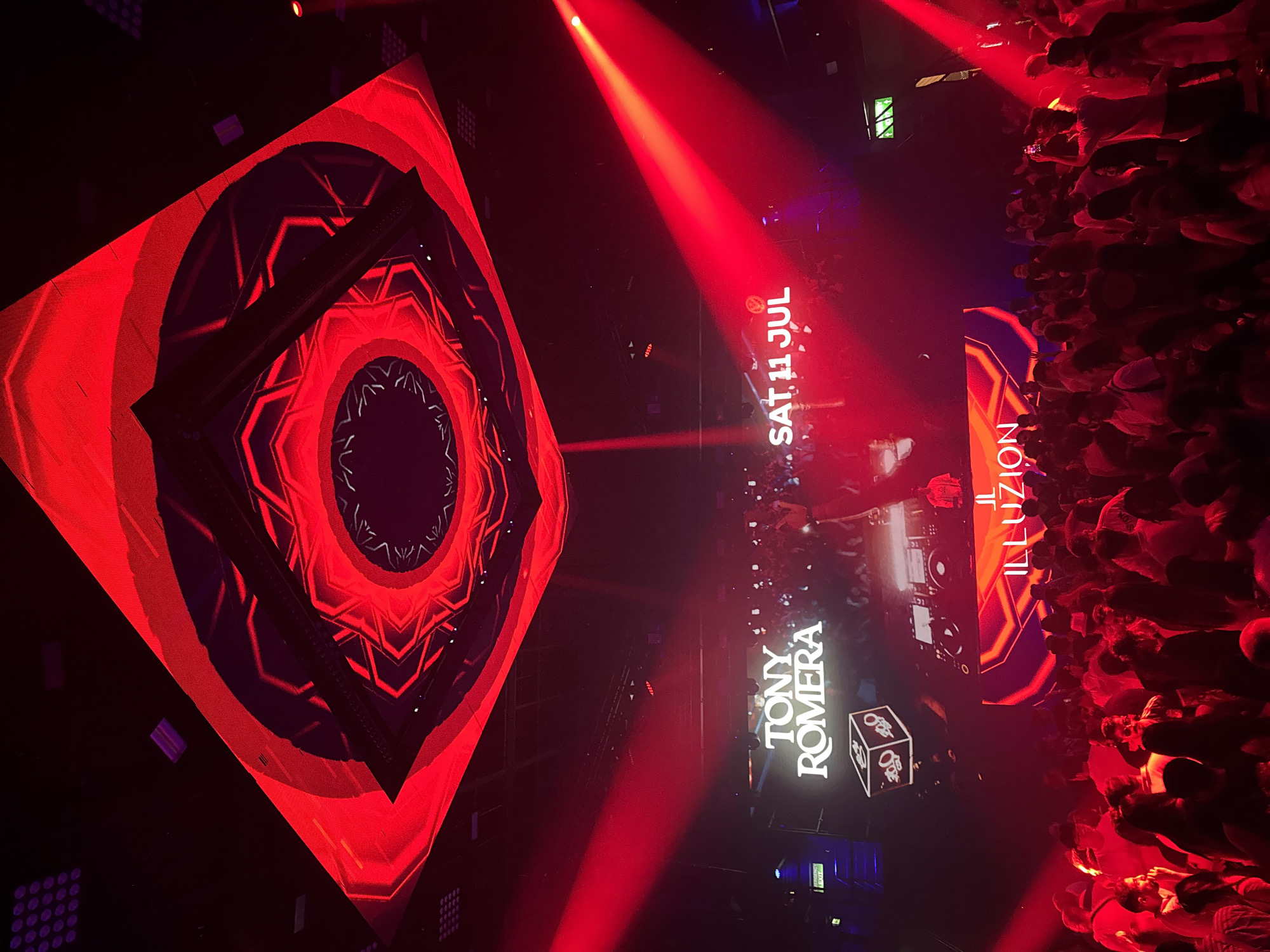 The image captures a vibrant nightclub scene in Patong, Thailand, dominated by intense red lighting. In the background, a large LED screen displays the text "TONY ROMERA" and "SAT 11 JUL," indicating a performance or event. Above the screen, a circular, geometric light installation glows red and purple. In the foreground, a dense crowd of people are visible, many holding up their phones, suggesting they are recording or taking pictures. The atmosphere is energetic, with red spotlights creating dramatic beams across the venue.