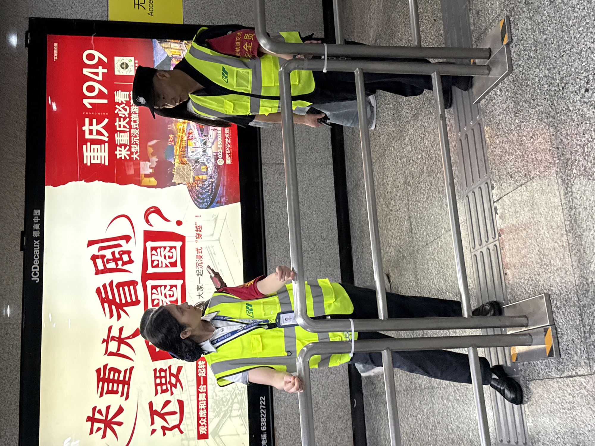 Two people wearing yellow safety vests stand on either side of metal handrails. Behind them is an advertisement for "Chongqing 1949," a large immersive tourist production, emphasizing the slogan "Come to Chongqing and watch the drama, or else?" in Yuzhong District, China.