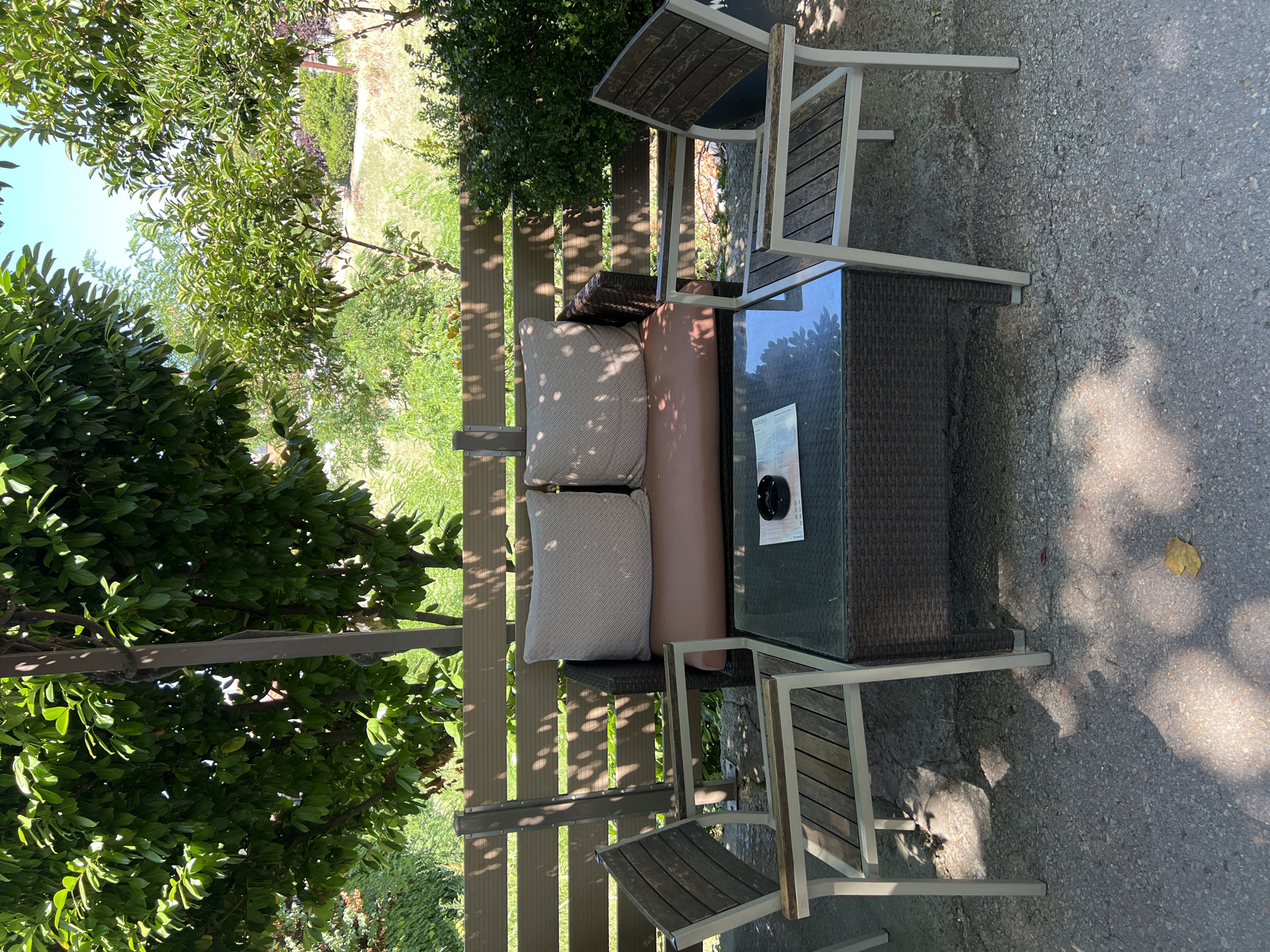 A shaded outdoor seating area in a city, possibly a cafe or restaurant, in Prokopio, Greece. The scene features a brown wicker loveseat with two textured light-colored cushions, accompanied by two matching chairs with wooden slats and light-colored metal frames. A square glass-topped coffee table made of brown wicker sits between the seating, holding a small black ashtray and a folded paper, possibly a menu. The furniture is situated on a gray asphalt or concrete ground, dappled with sunlight and shadows filtering through the dense green foliage of trees overhead. A wooden slatted fence forms the backdrop, behind which more greenery and a slightly sloped, dry grassy area can be seen. The environment suggests a warm, sunny day.