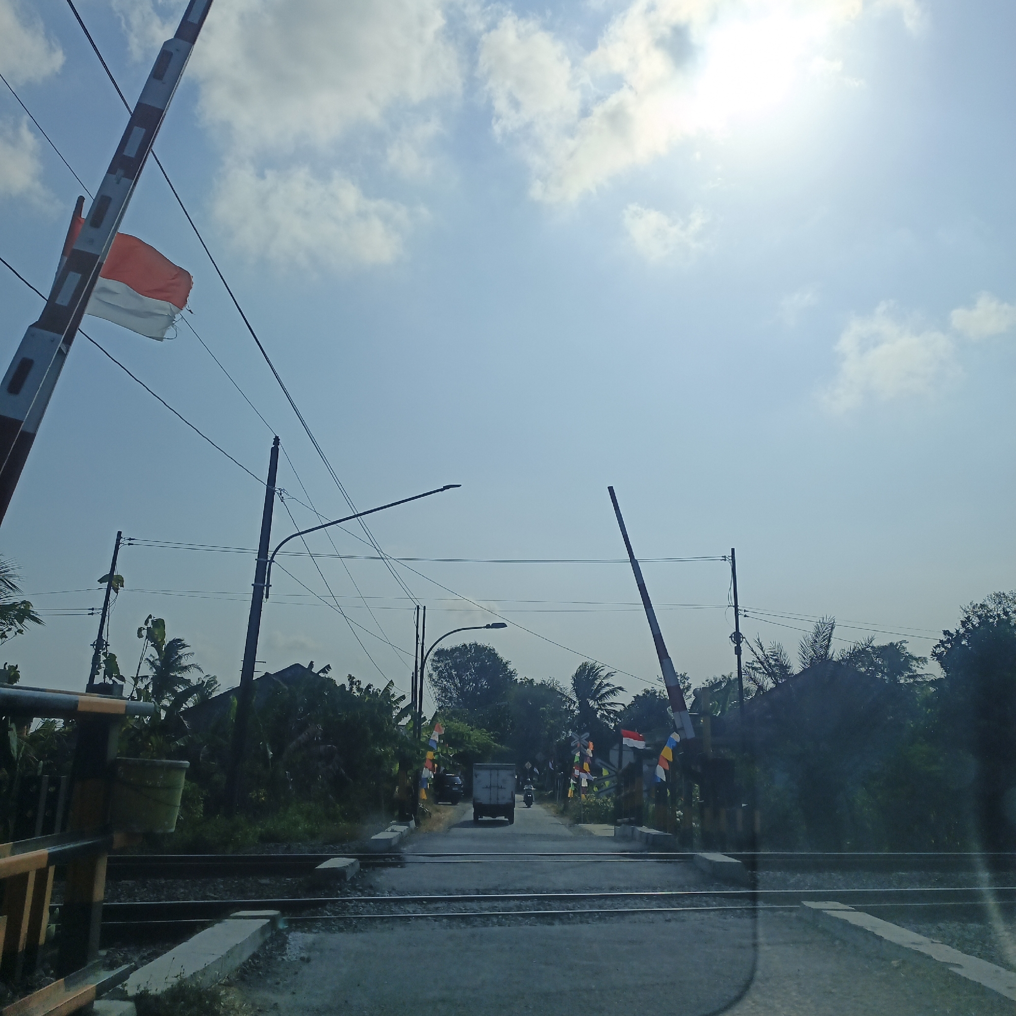 The image depicts a railway crossing in Bagelen, Indonesia, under a bright, sunny sky with scattered clouds. A red and white striped railway barrier arm is raised on the left side, adorned with an Indonesian flag. Another barrier arm is raised on the right side. The road leading up to the crossing has several small flags lining its sides. In the distance, a truck and a motorcycle are visible on the road. The environment is characterized by lush green vegetation and trees on either side of the road. Power lines and street lamps are also present. The railway tracks run horizontally across the foreground.