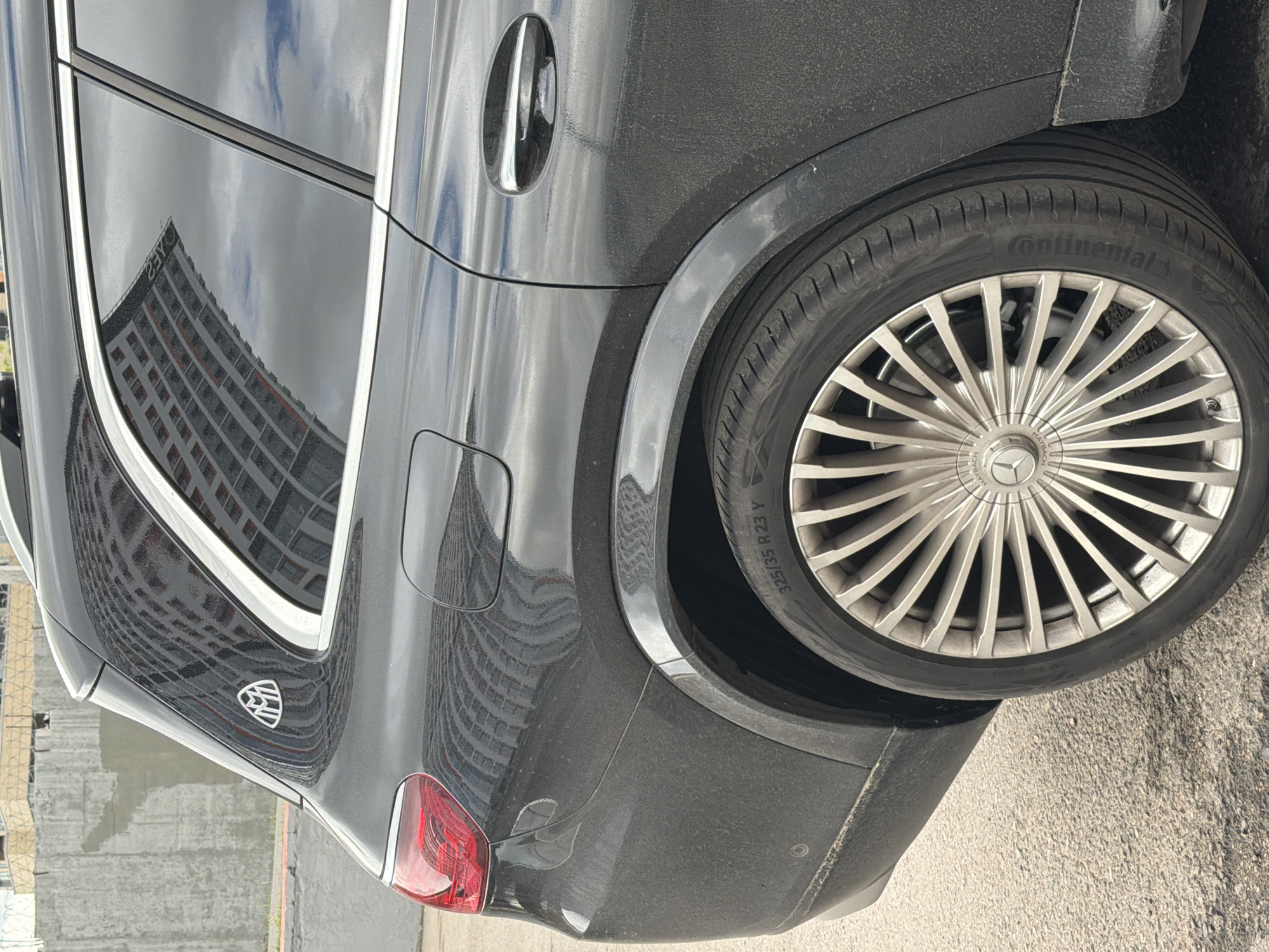 The image shows the rear of a black Maybach car, specifically the passenger side rear quarter panel, wheel, and taillight. The tire brand is Continental, and the tire size is 325/35 R23. The car's reflection shows a building, which identifies the location as Moscow, Russia. The car is parked on asphalt and appears to be dirty.