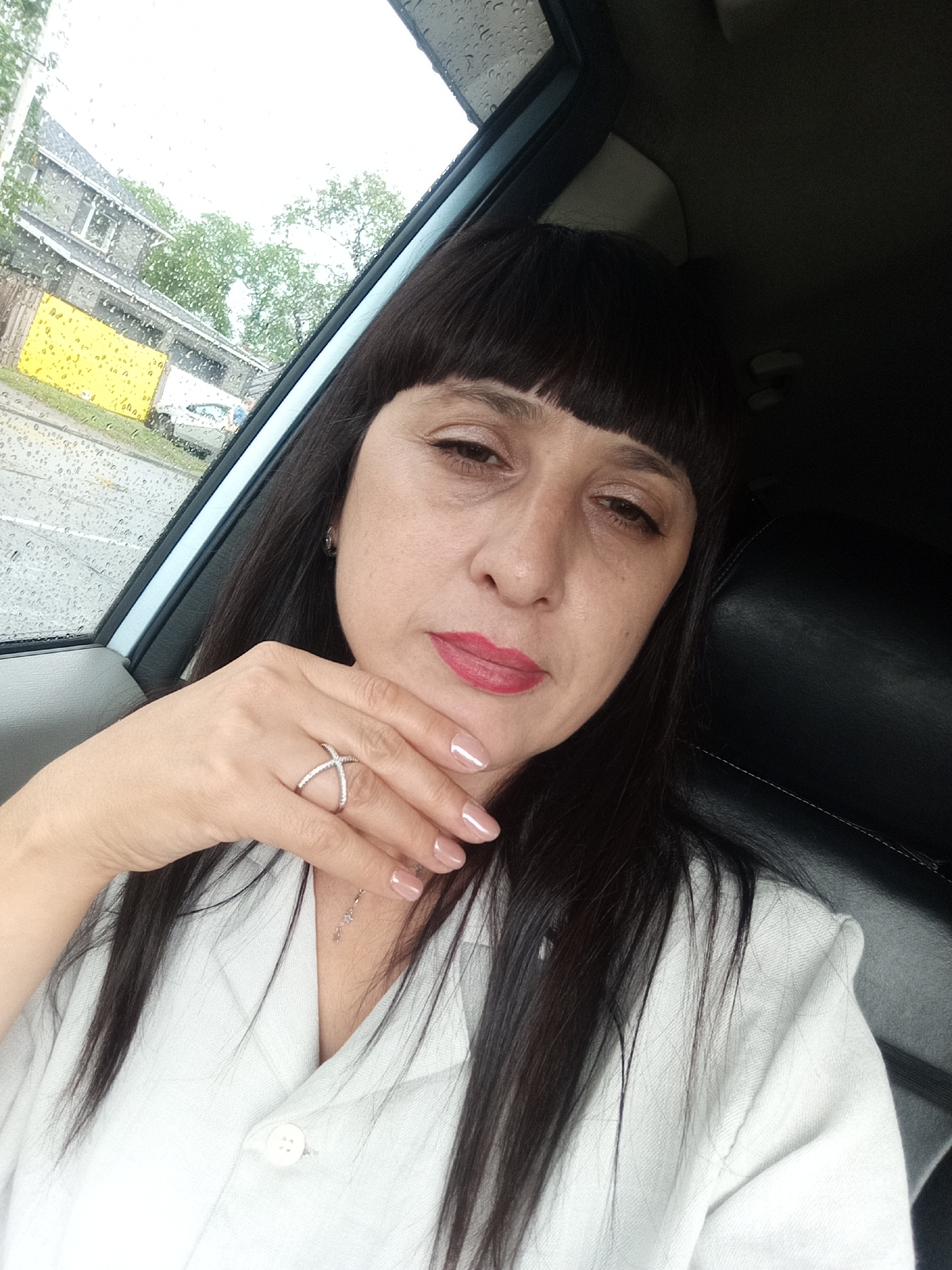In a selfie taken from inside a vehicle, a woman with dark hair and bangs rests her chin on her hand, adorned with a sparkly criss-cross ring. She is wearing a light-colored collared shirt. Raindrops are visible on the car window, obscuring the view of a street with buildings, trees, and a yellow structure. The scene appears to be in the city of Belogorsk, Russia.