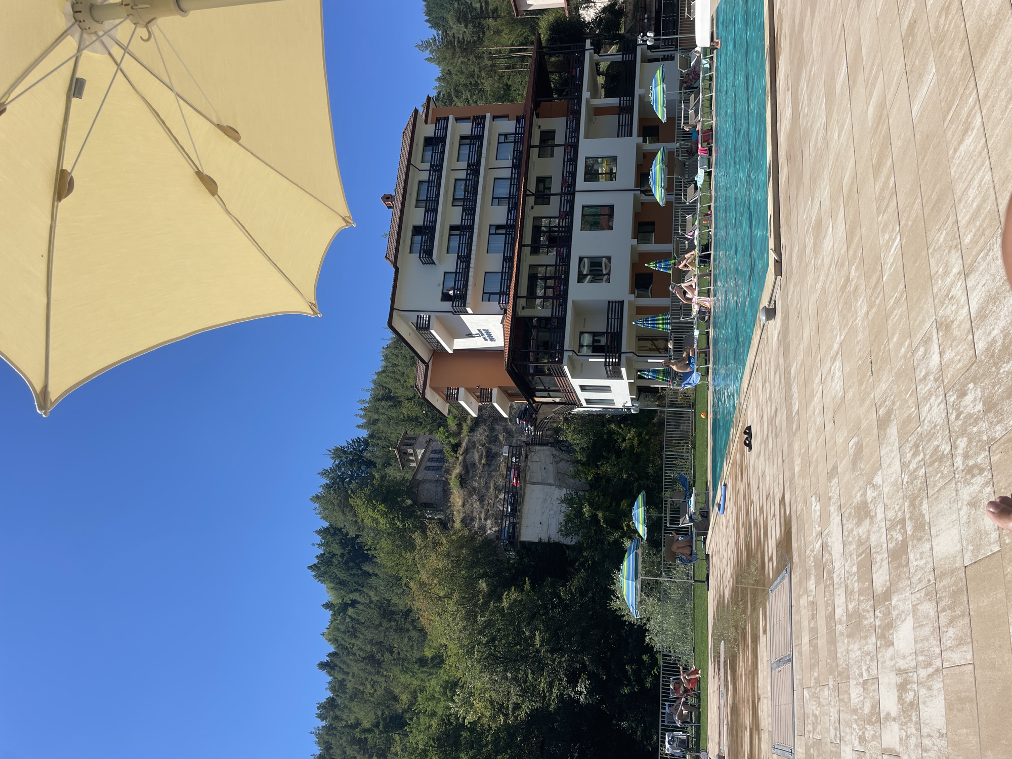 The image shows a swimming pool area with people relaxing on lounge chairs. A multi-story building with balconies is visible in the background, with a forested hill behind it. There is also a small building on a cliff face. The sky is clear and blue. The scene is outdoors on a sunny day. The building in the background is the Hotel "Prilep" in the town of Banite, Bulgaria, known for its mineral springs.